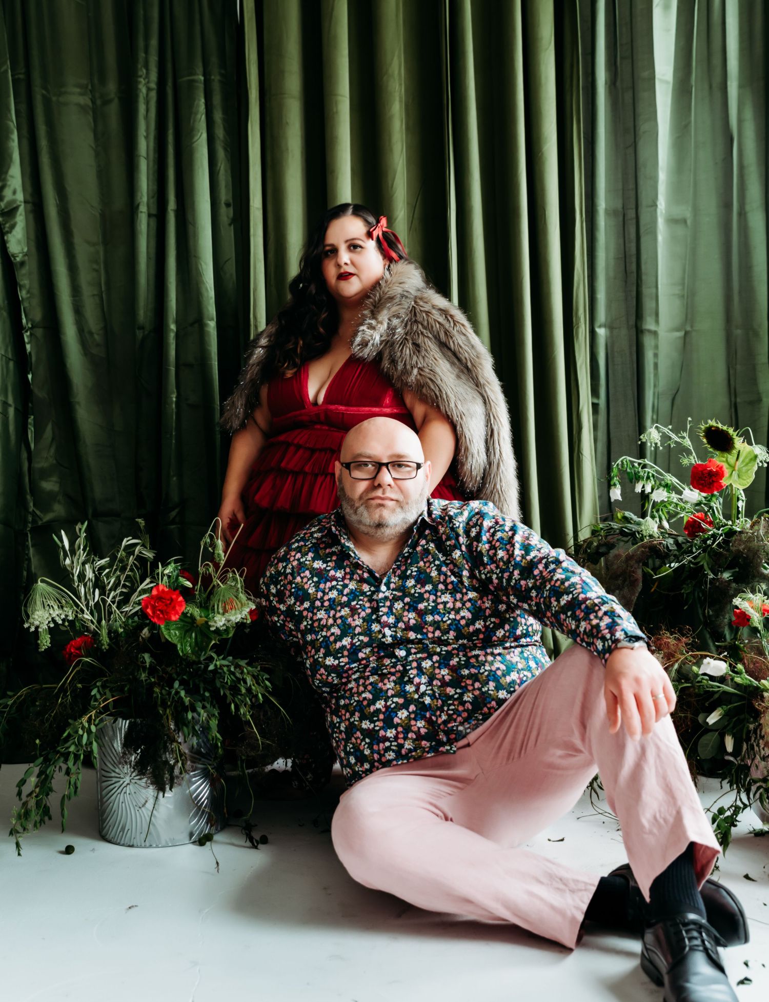 A figure in a floral print top and pink pants reclines against a green curtain backdrop with potted red flowers.