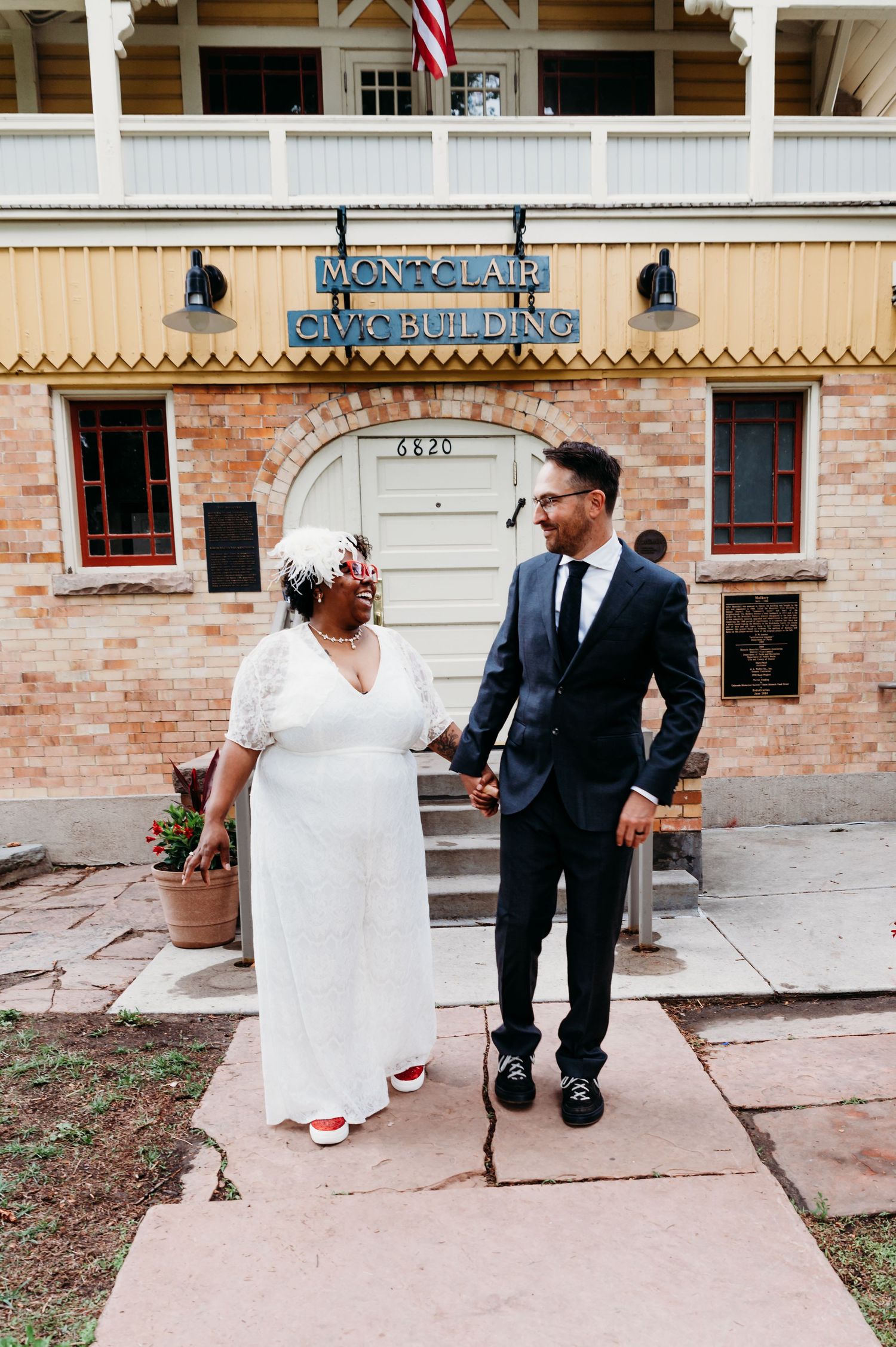 A couple in elegant wedding attire poses in front of a brick building with a 'Manvalley' sign.