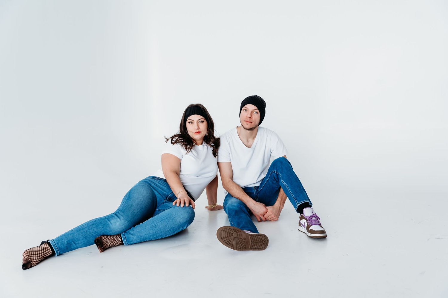 A casual couple in matching white t-shirts and jeans sits together on a white studio floor.