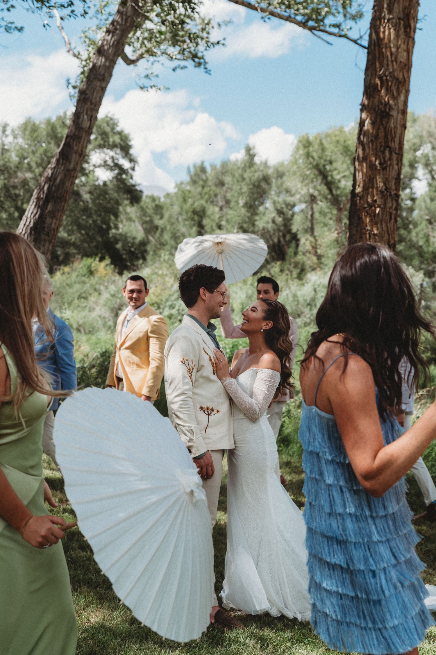 Bride and groom share a kiss surrounded by guests holding white parasols at an outdoor wedding ceremony.