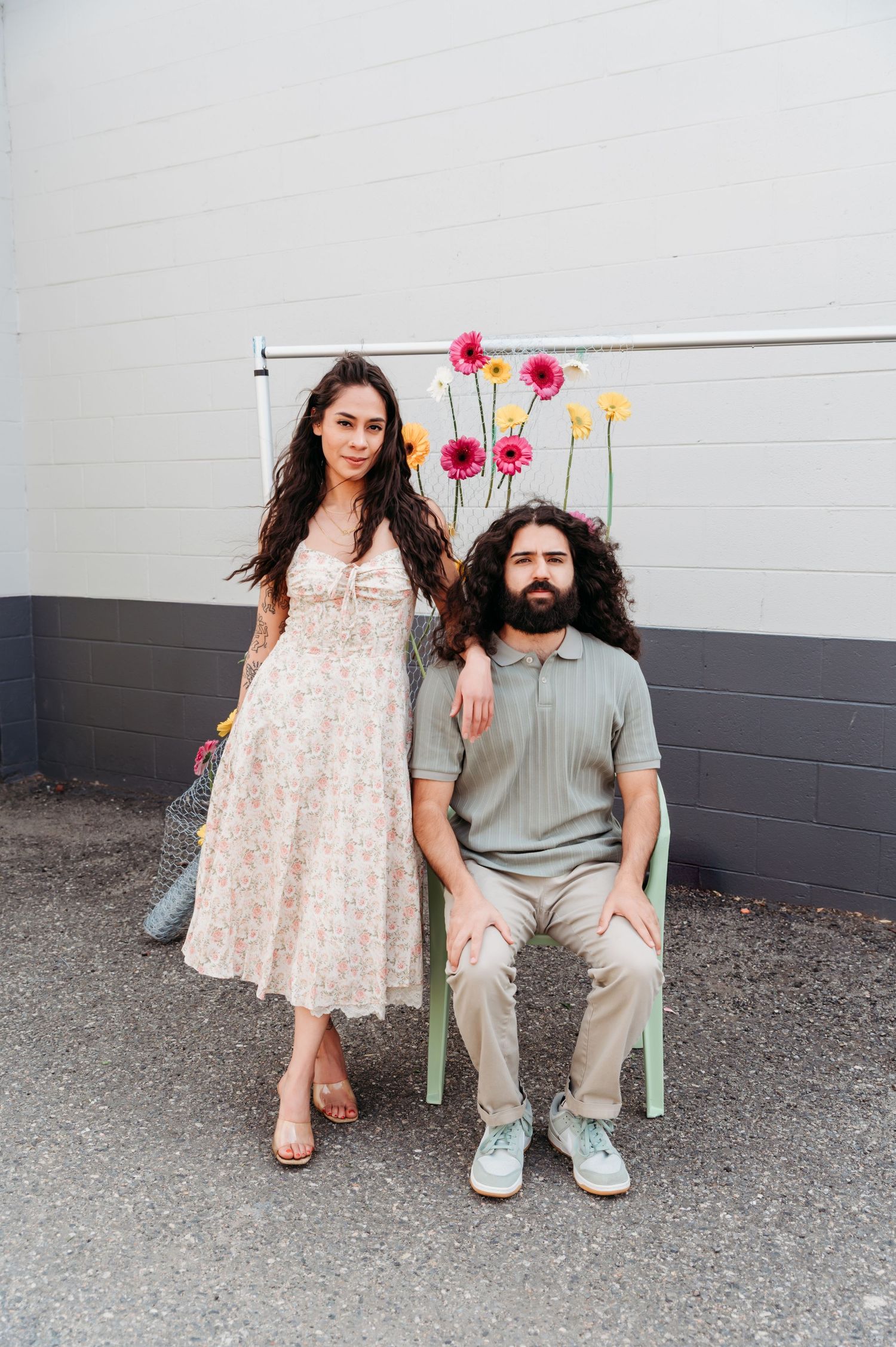 A couple sits together on a bench against a gray wall while holding pink flowers in a casual outdoor setting.