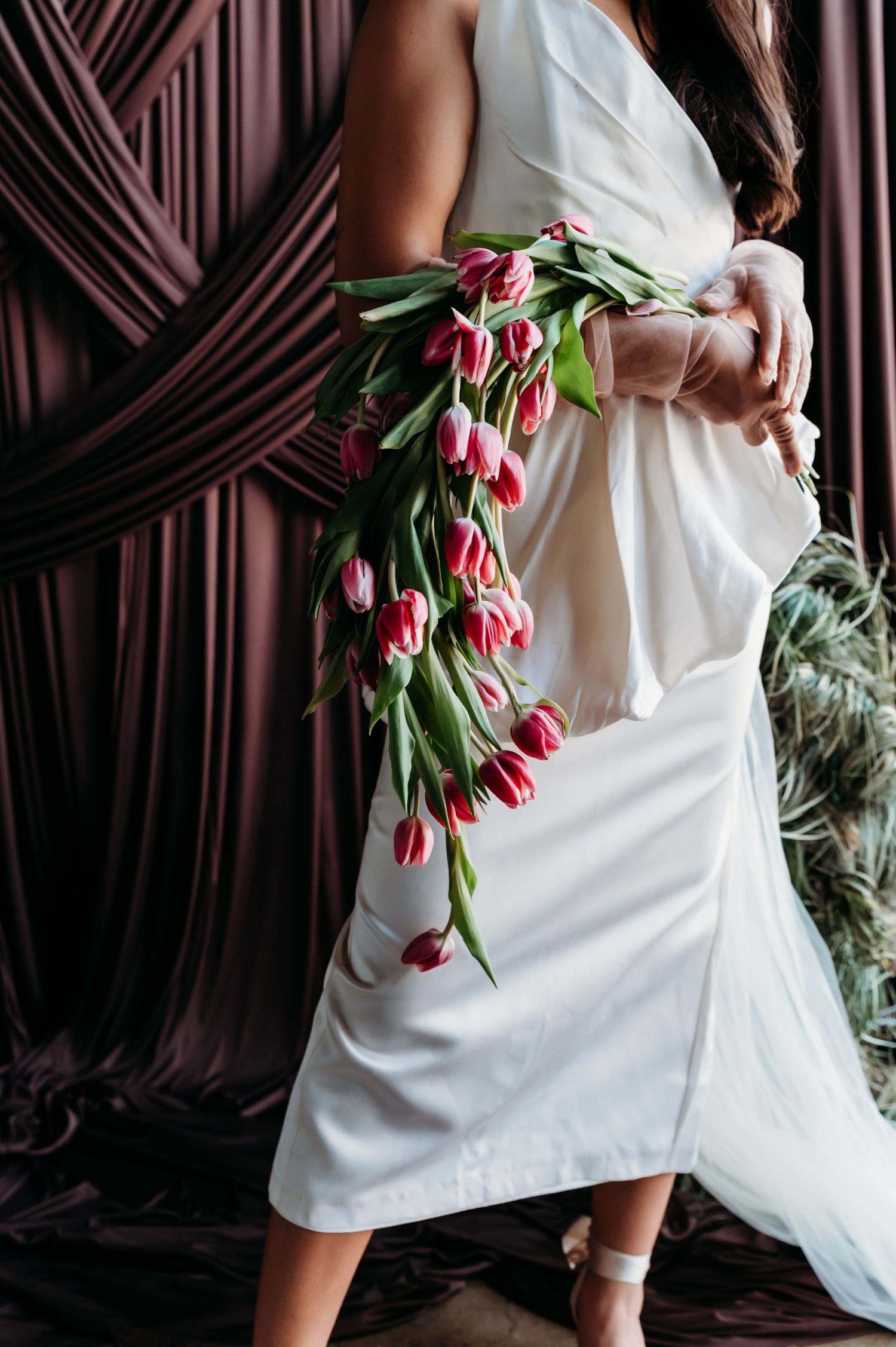 Woman in white dress holding cascading red tulips bouquet against mauve draped curtain backdrop.