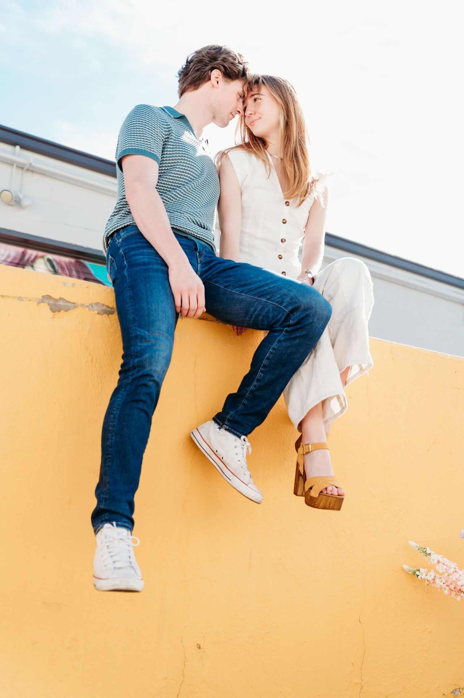 Couple sitting intimately on a yellow wall in casual denim and white outfits.