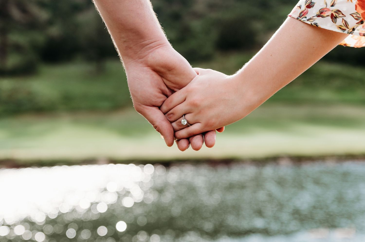 Two hands gently clasped together against a sunlit water backdrop in a tender moment of connection.