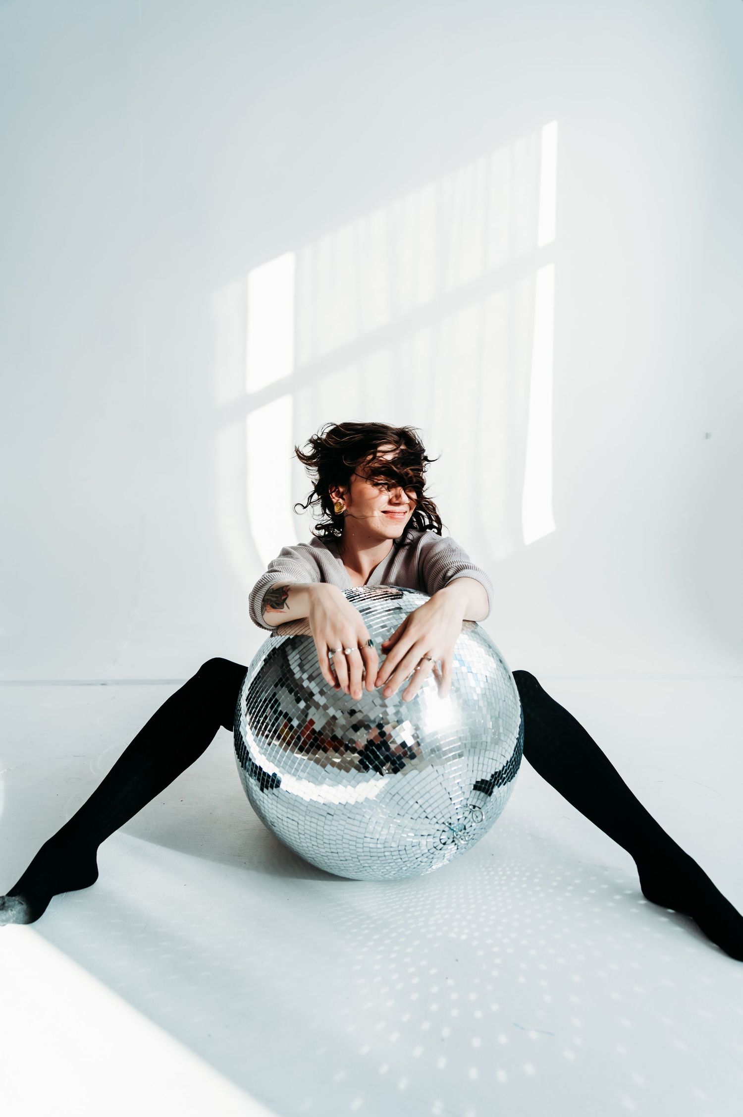 A person poses with a large disco ball in a minimalist white room.