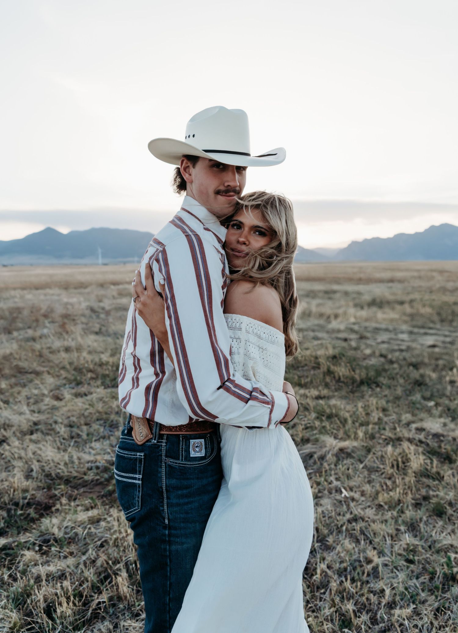 A couple in western attire embraces in a field at sunset with mountains in the background.