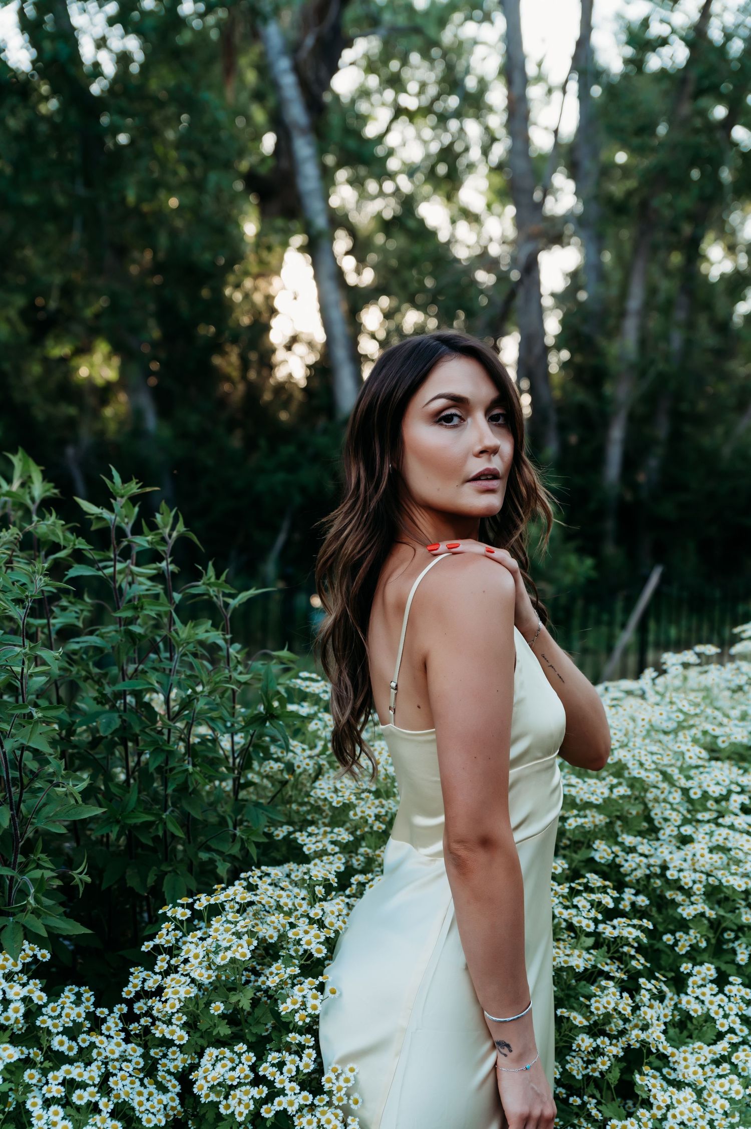 A person in a cream-colored dress poses among blooming white flowers and greenery in a forest setting at dusk.