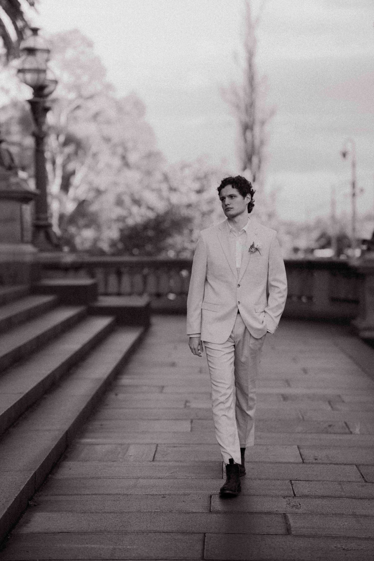 Groom in cream suit walking on stone steps at outdoor wedding venue in black and white photo.