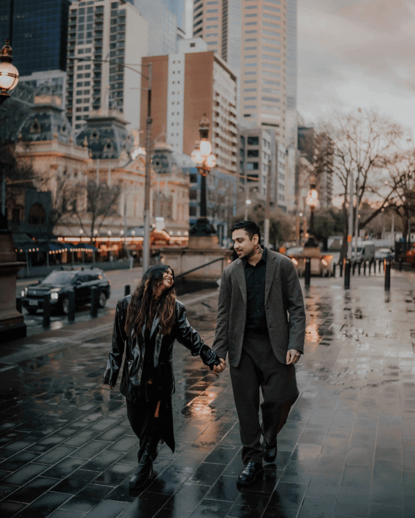 Couple holds hands walking on rain-soaked city street at dusk, surrounded by glowing lights and urban architecture.