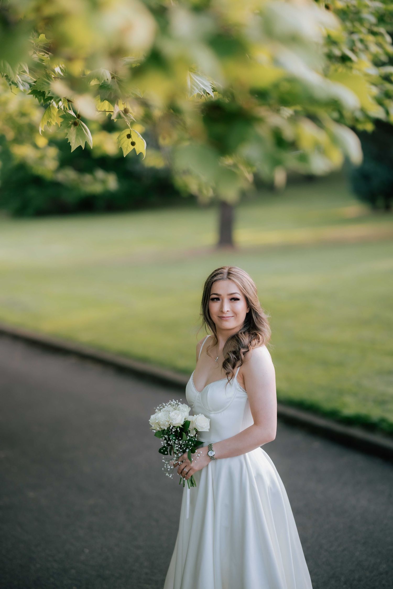 Bride in a white satin gown holding a white bouquet while standing on a tree-lined path.