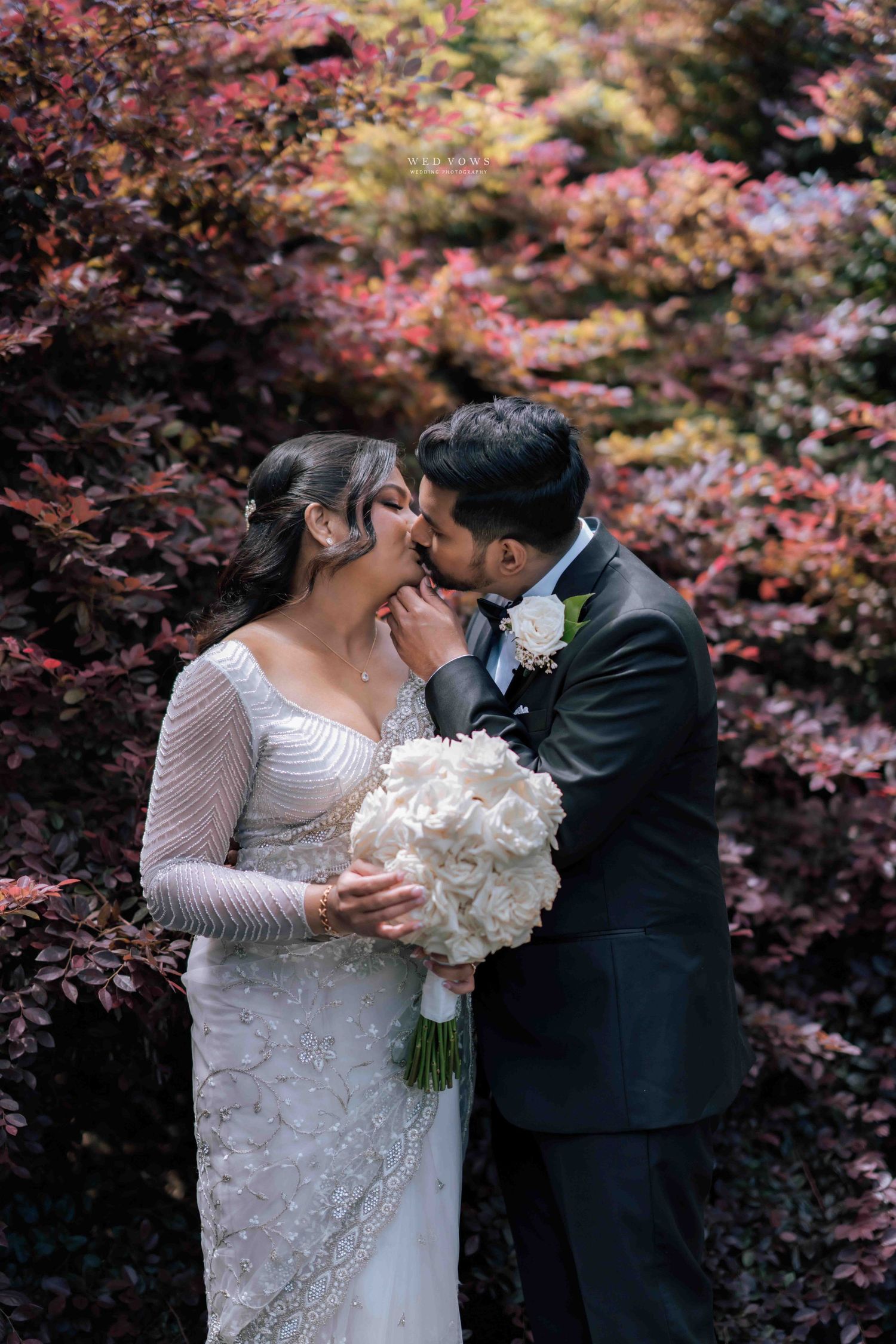 Bride and groom kissing in front of vibrant red autumn foliage, bride holding white hydrangea bouquet.