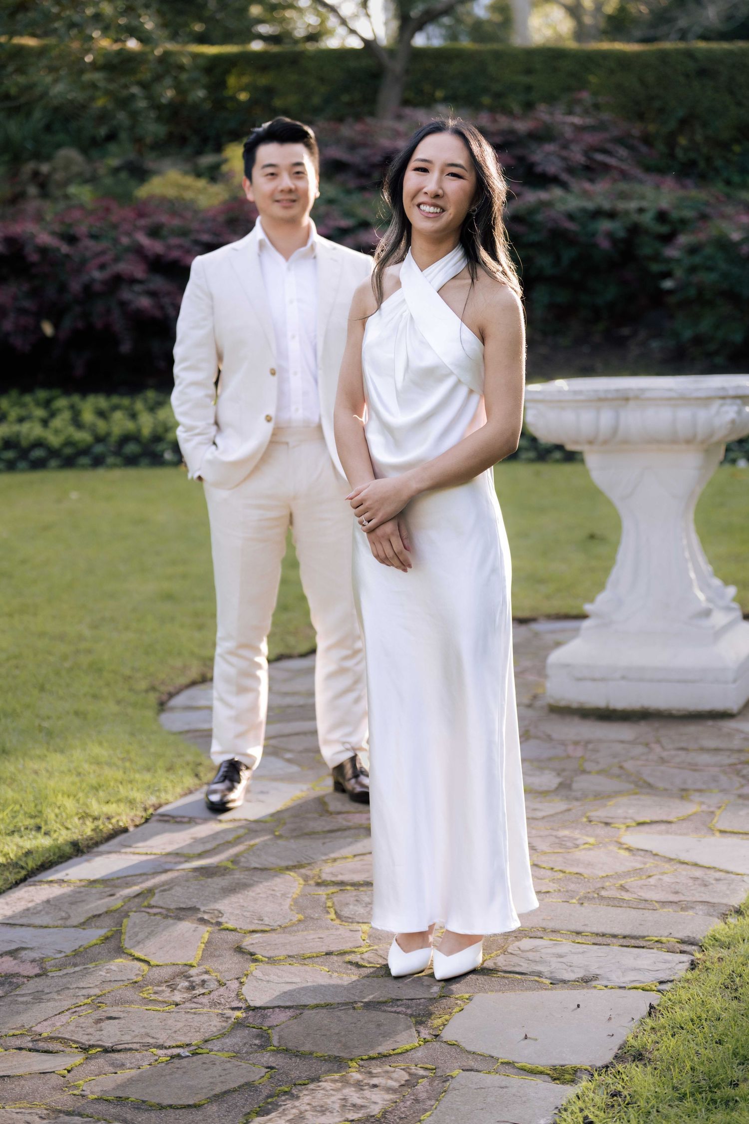 Bride in white halter dress and groom in cream suit posing on stone garden path with lush greenery backdrop.