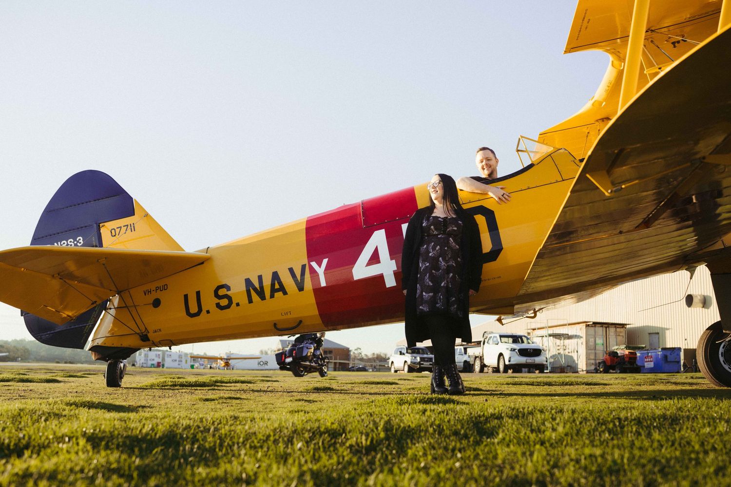 Couple poses beside a vintage U.S. Navy yellow biplane on a grass airfield at sunset.