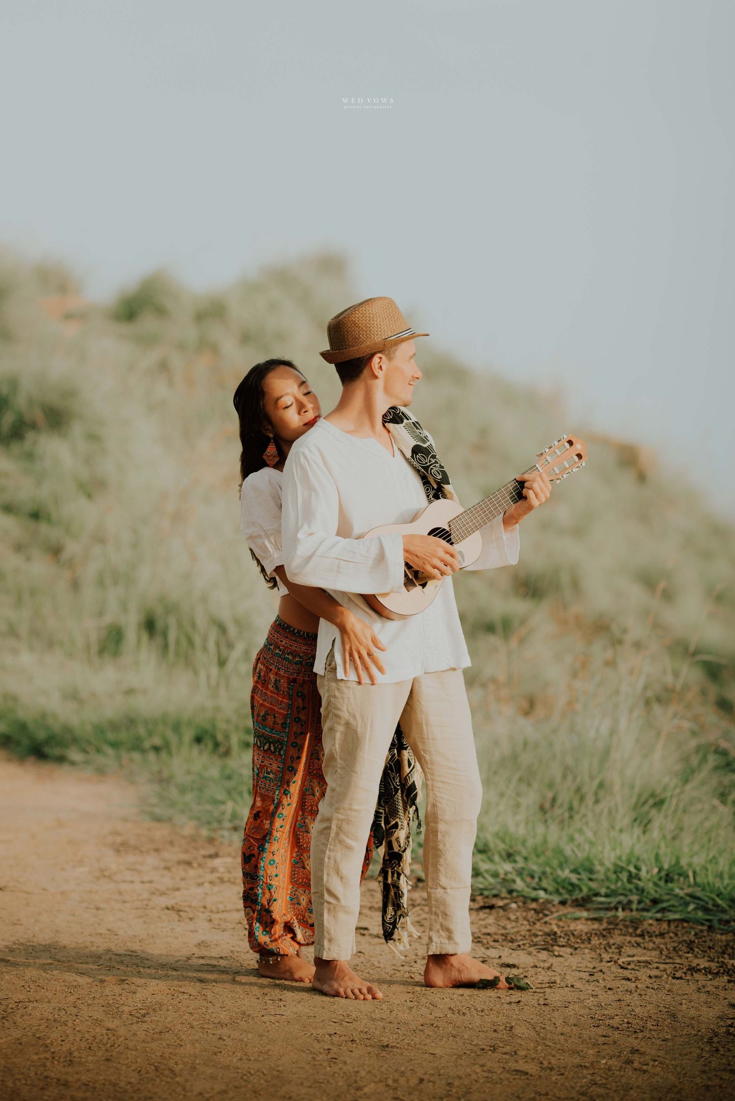 Woman embraces man playing ukulele on sunny hillside path wearing straw hat.