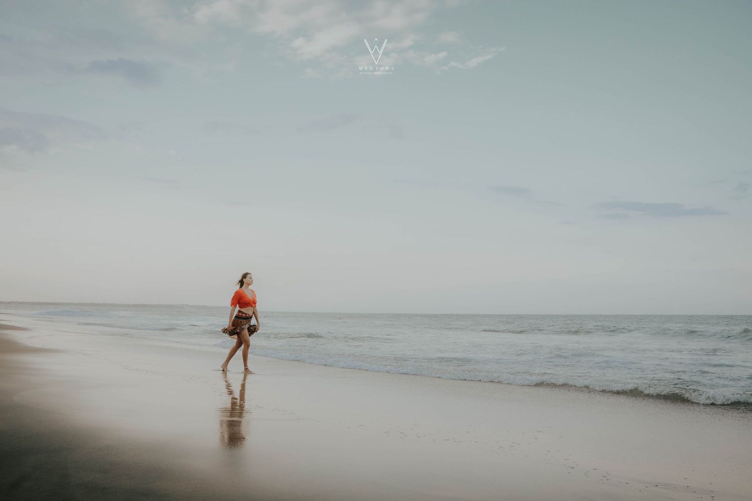 Woman in orange crop top walking alone along a calm tropical beach shoreline at dusk.