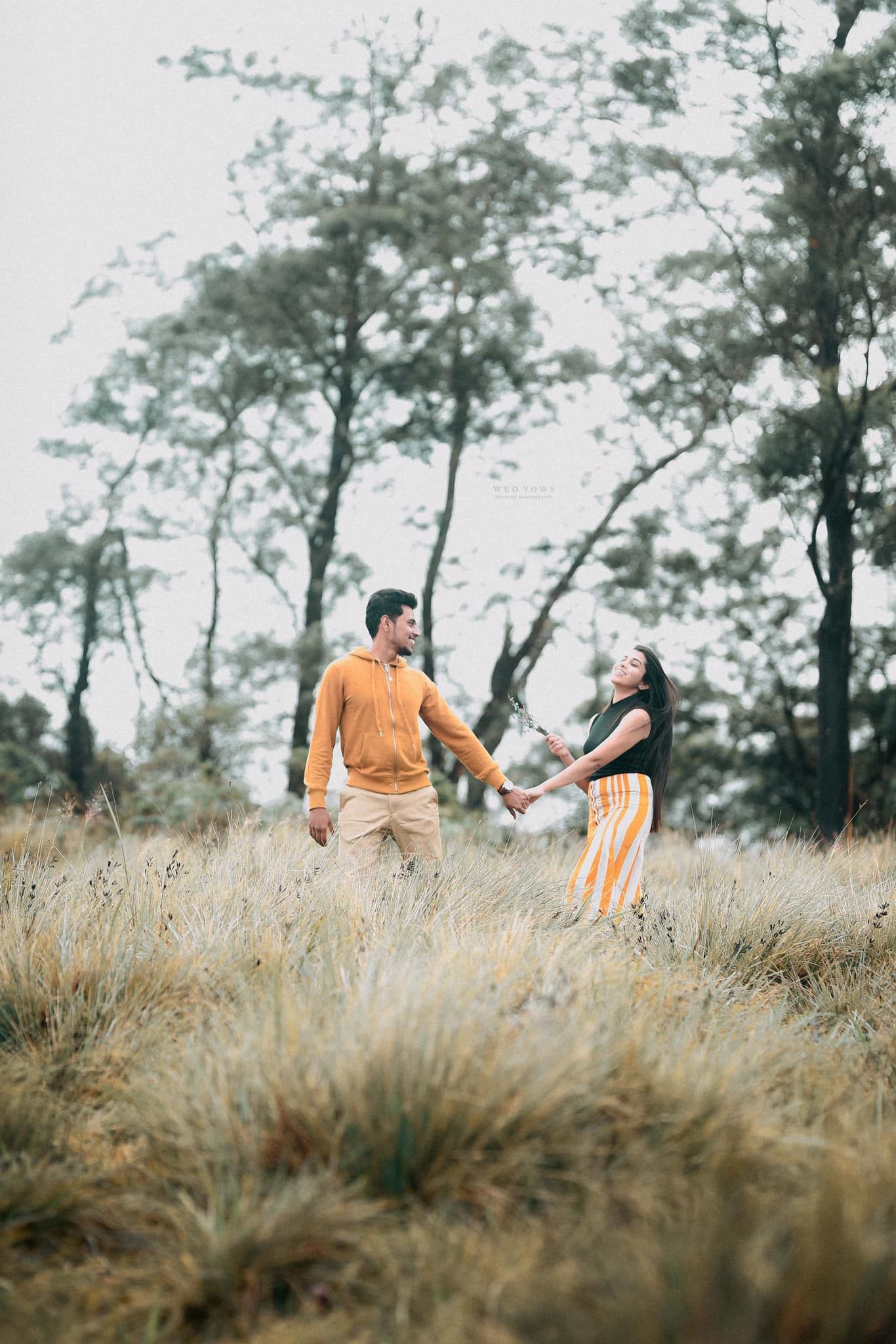 Couple holding hands in golden grass field with trees, woman looking up joyfully at the sky.
