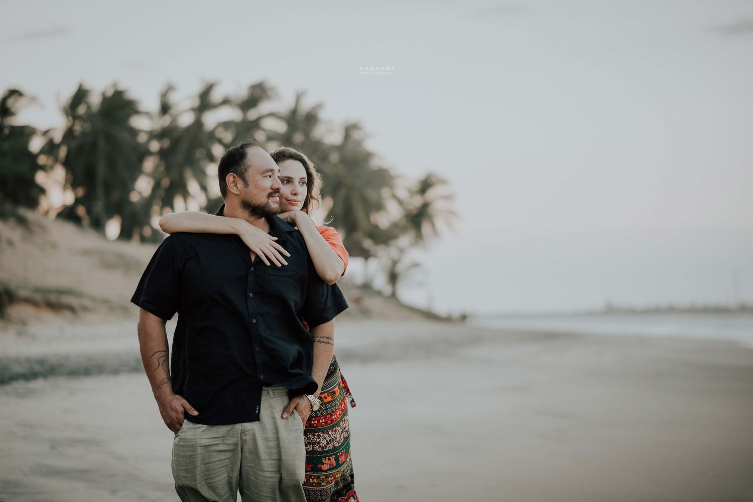 Couple embracing on a tropical beach with palm trees, woman hugging man from behind at dusk.