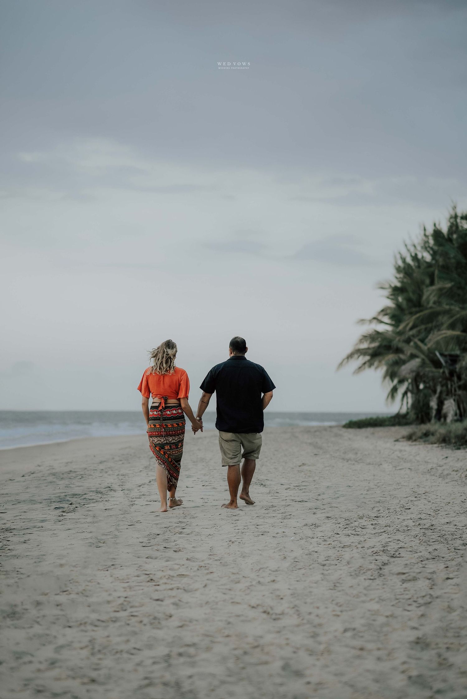 Couple walking hand in hand along sandy tropical beach with palm trees in distance.
