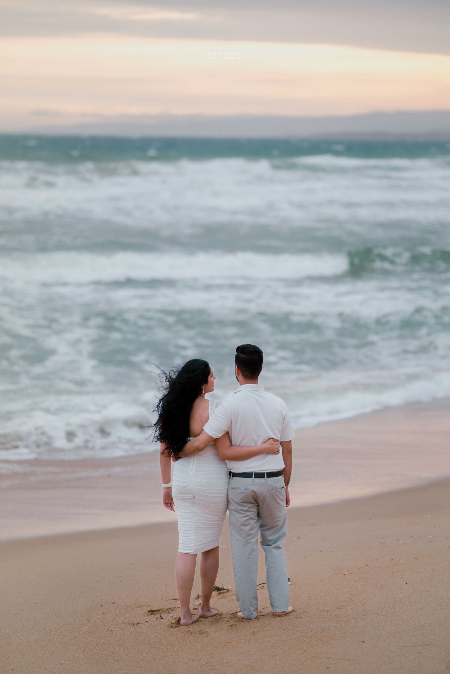 Couple in white outfits embrace while watching ocean waves at sunset on a sandy beach.