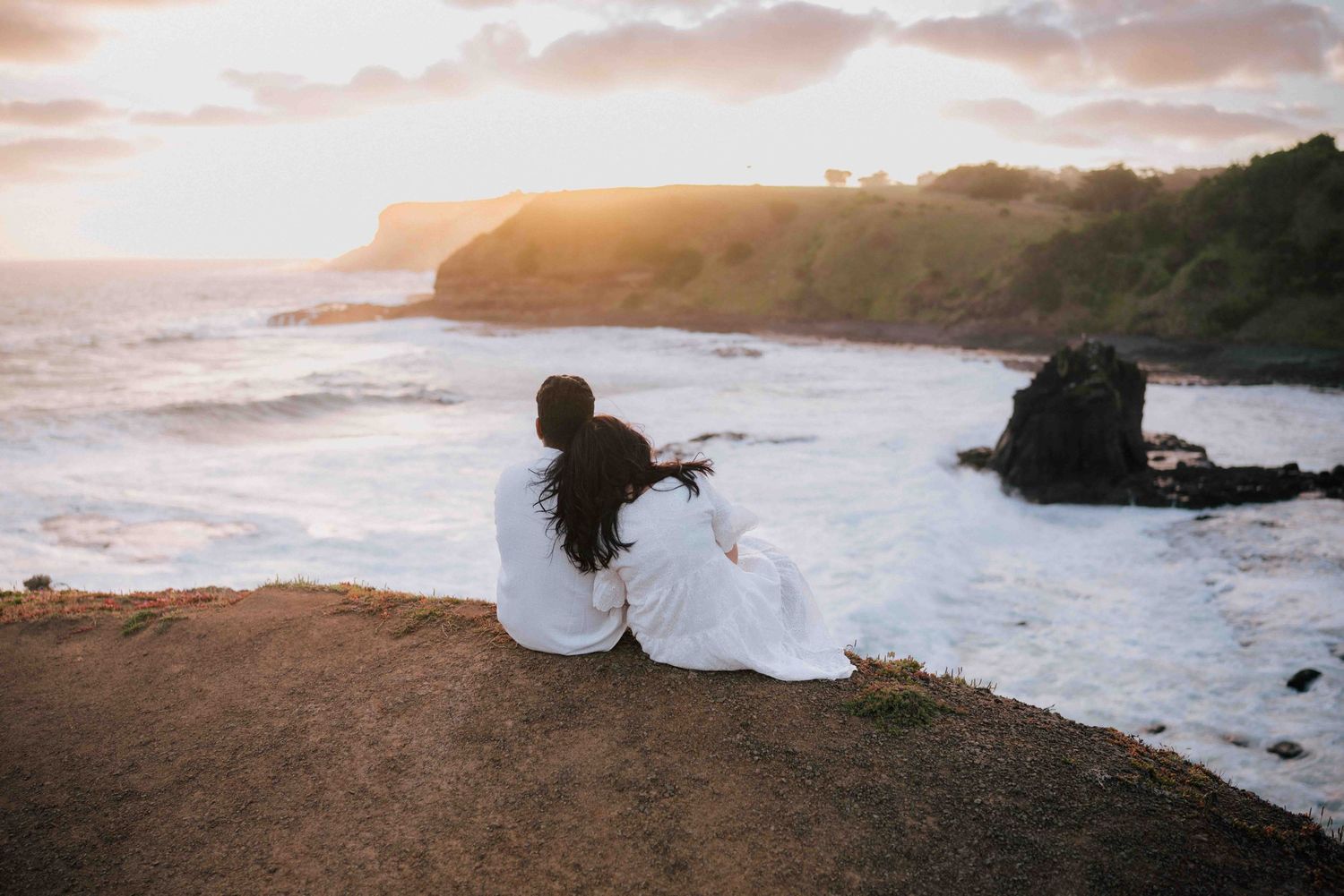 Couple sitting together on coastal cliff watching golden sunset over ocean waves.
