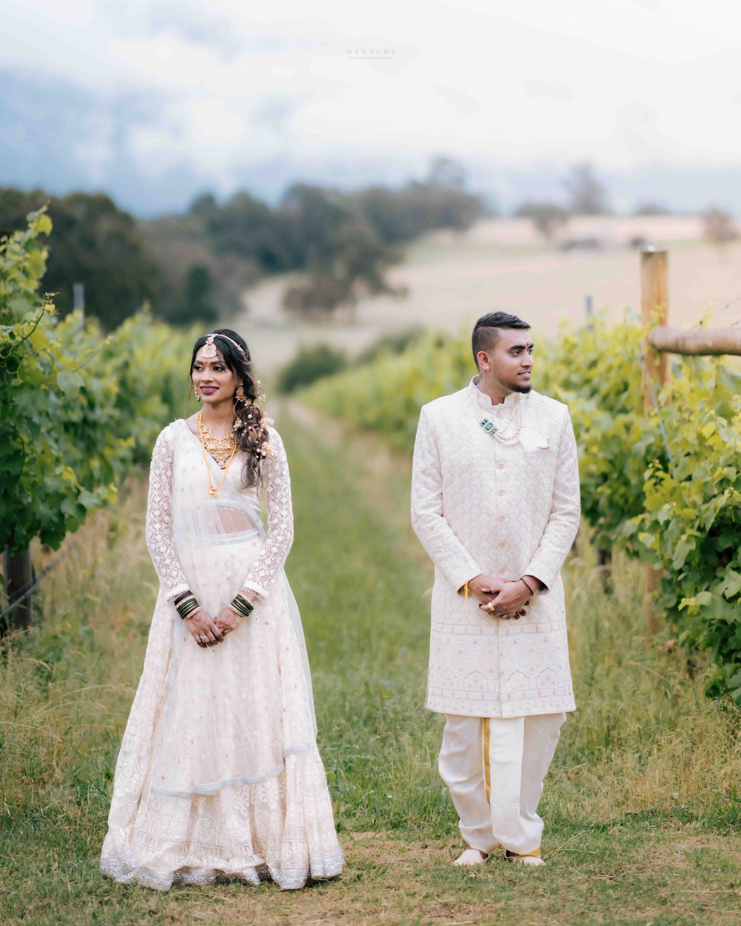 Indian bride and groom in matching white wedding outfits stand together between vineyard rows.