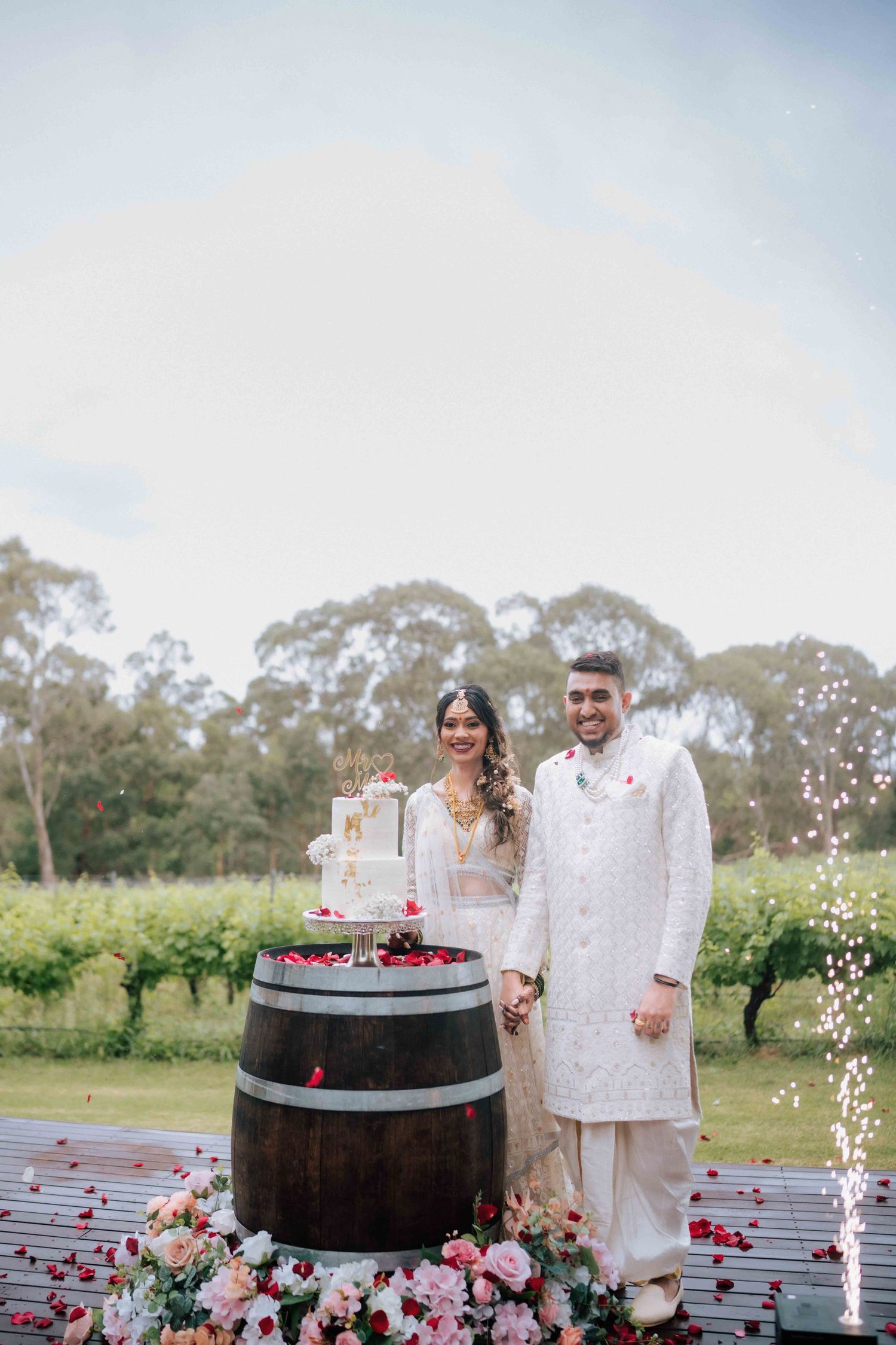 Indian wedding couple in white outfits standing beside tiered cake on barrel with floral arrangements.