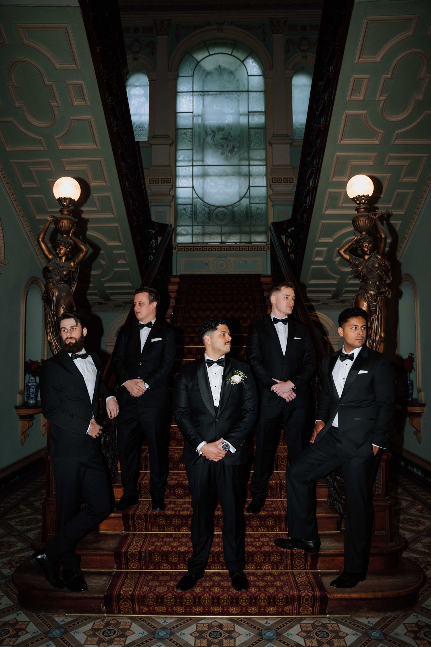 Groom and groomsmen in black tuxedos posing on ornate staircase with stained glass window backdrop.