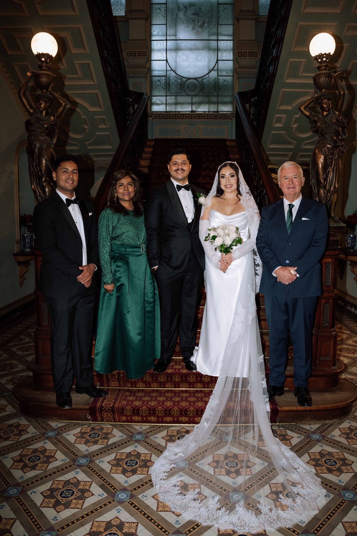 Wedding party family portrait on grand staircase, bride in white gown with cathedral veil holding white bouquet.