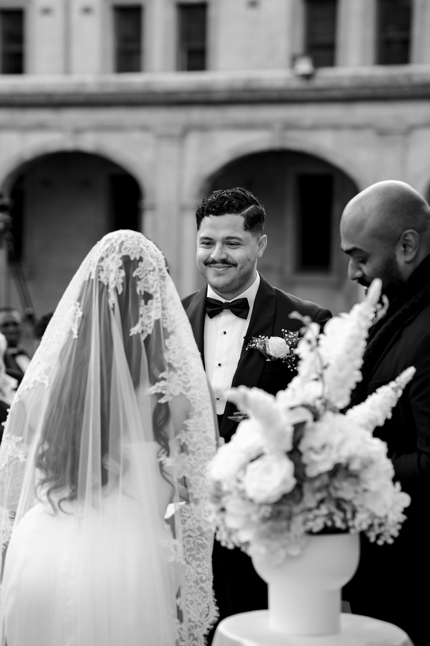 Black and white photo of groom smiling lovingly at veiled bride during outdoor wedding ceremony.