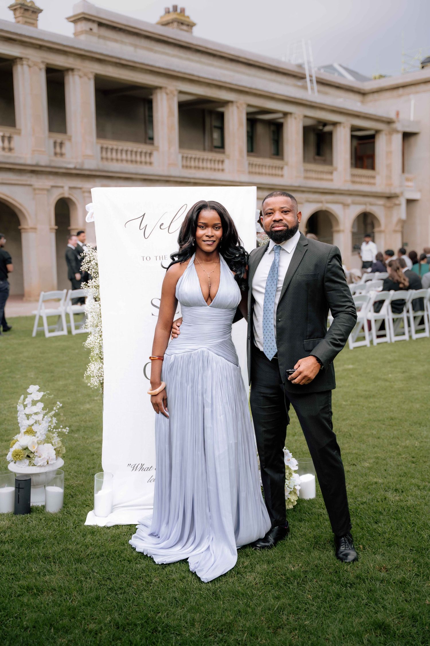 Couple posing in front of wedding welcome sign on lawn of elegant venue, woman in silver gown, man in dark suit.