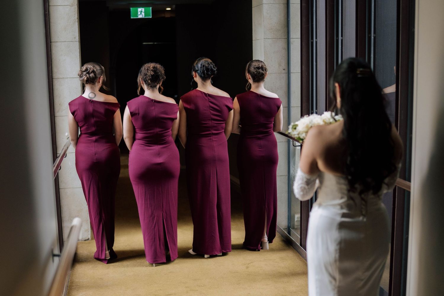 Bride with bouquet and four bridesmaids in burgundy gowns standing in hotel corridor.