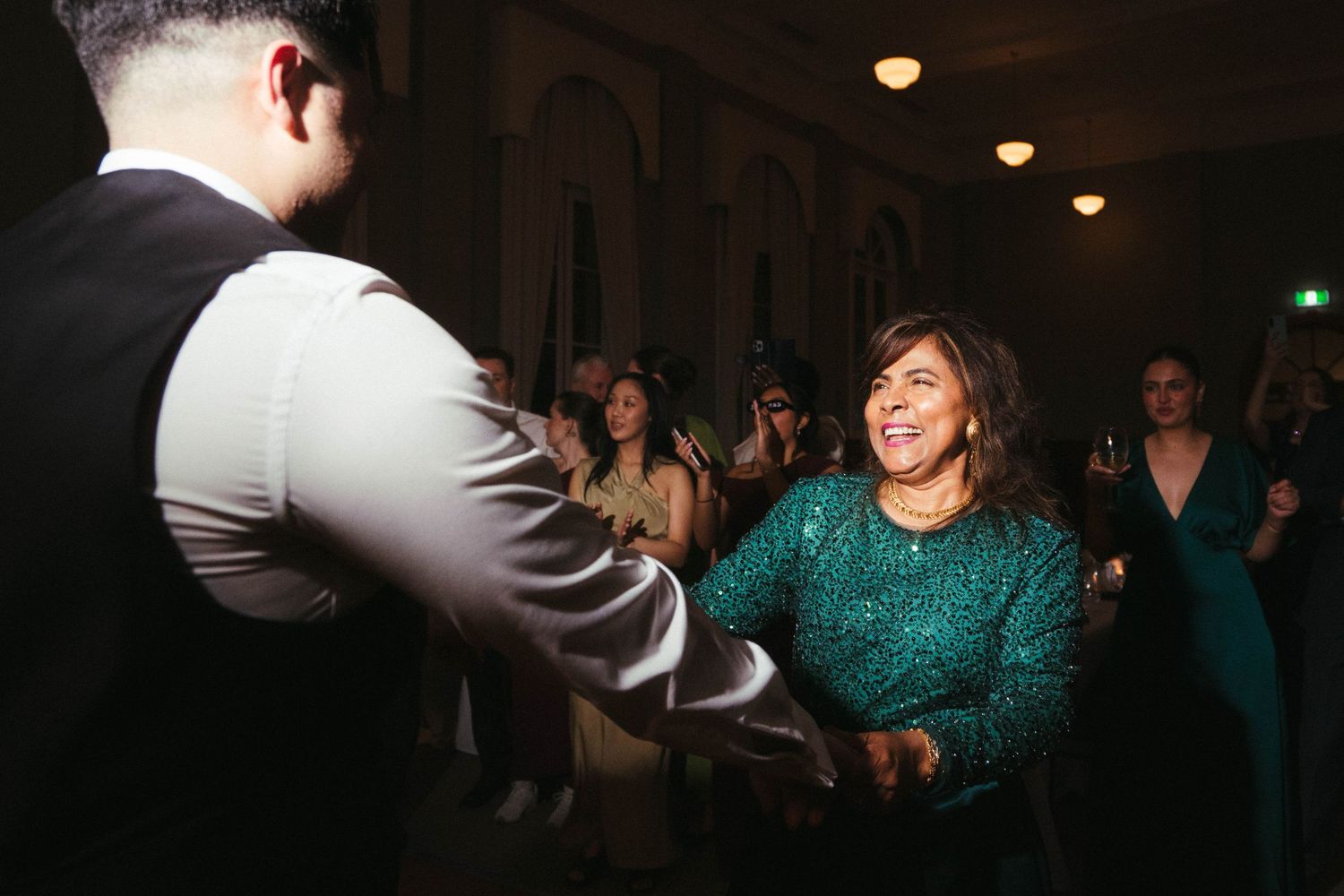 Groom shares a dance with smiling woman in green sequin dress at wedding reception.