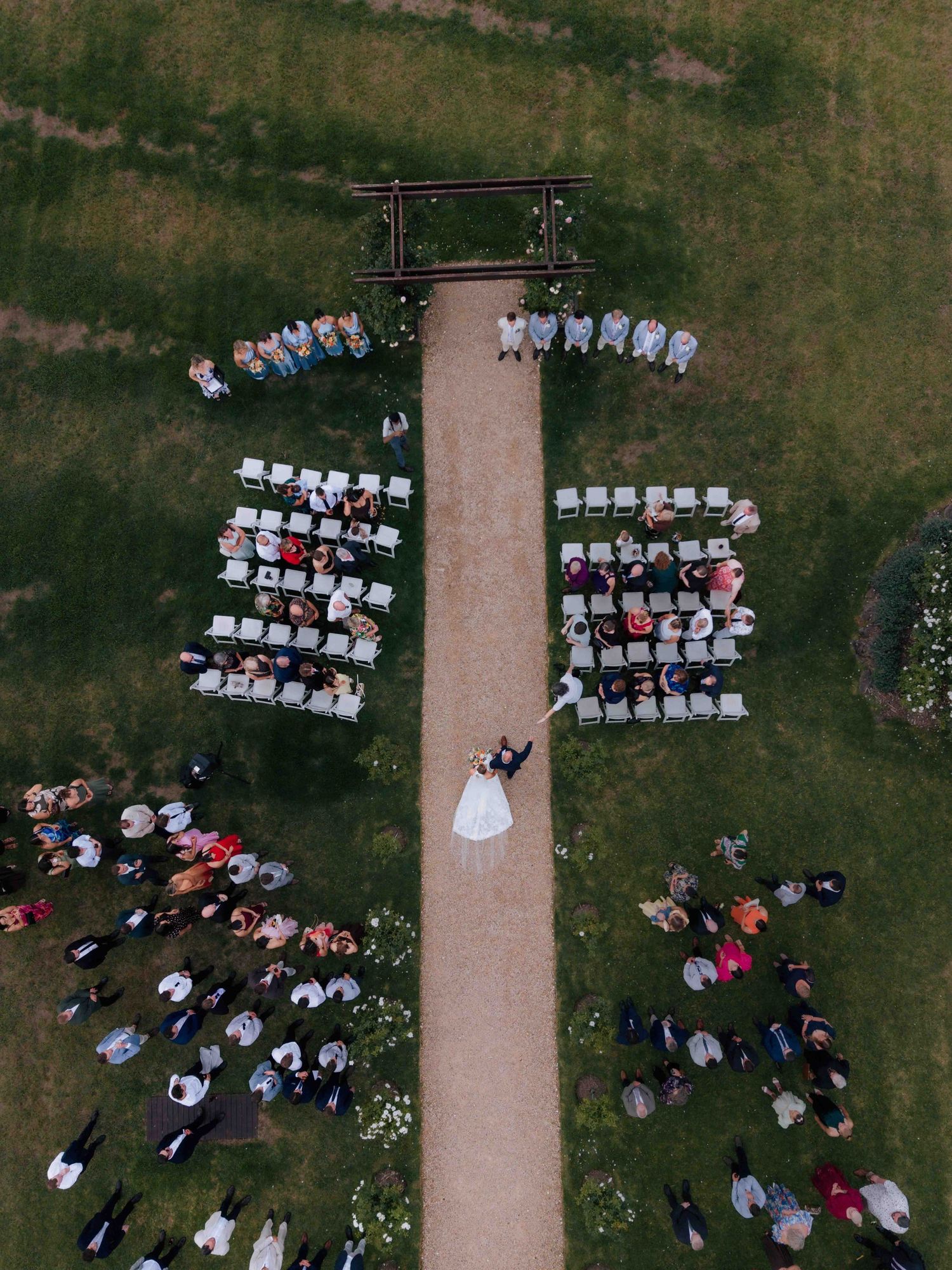 Aerial drone view of an outdoor wedding ceremony with guests seated in white chairs along a gravel aisle.