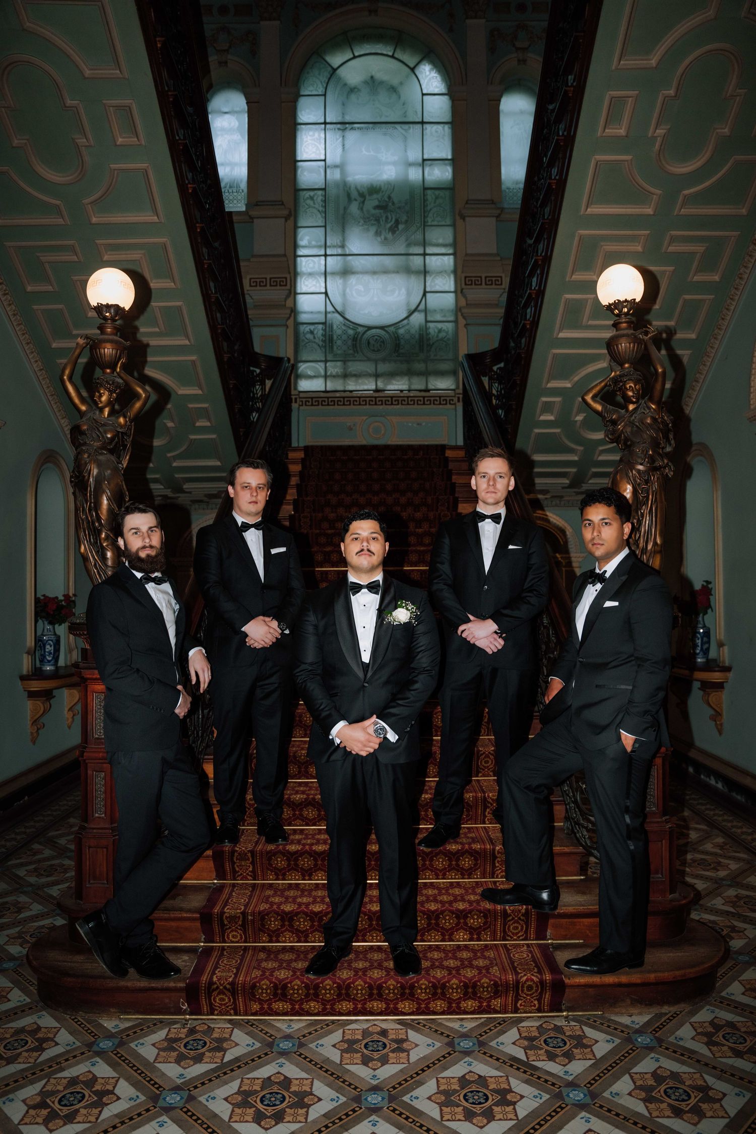Five groomsmen in black tuxedos pose on an ornate staircase with stained glass windows and bronze statues.
