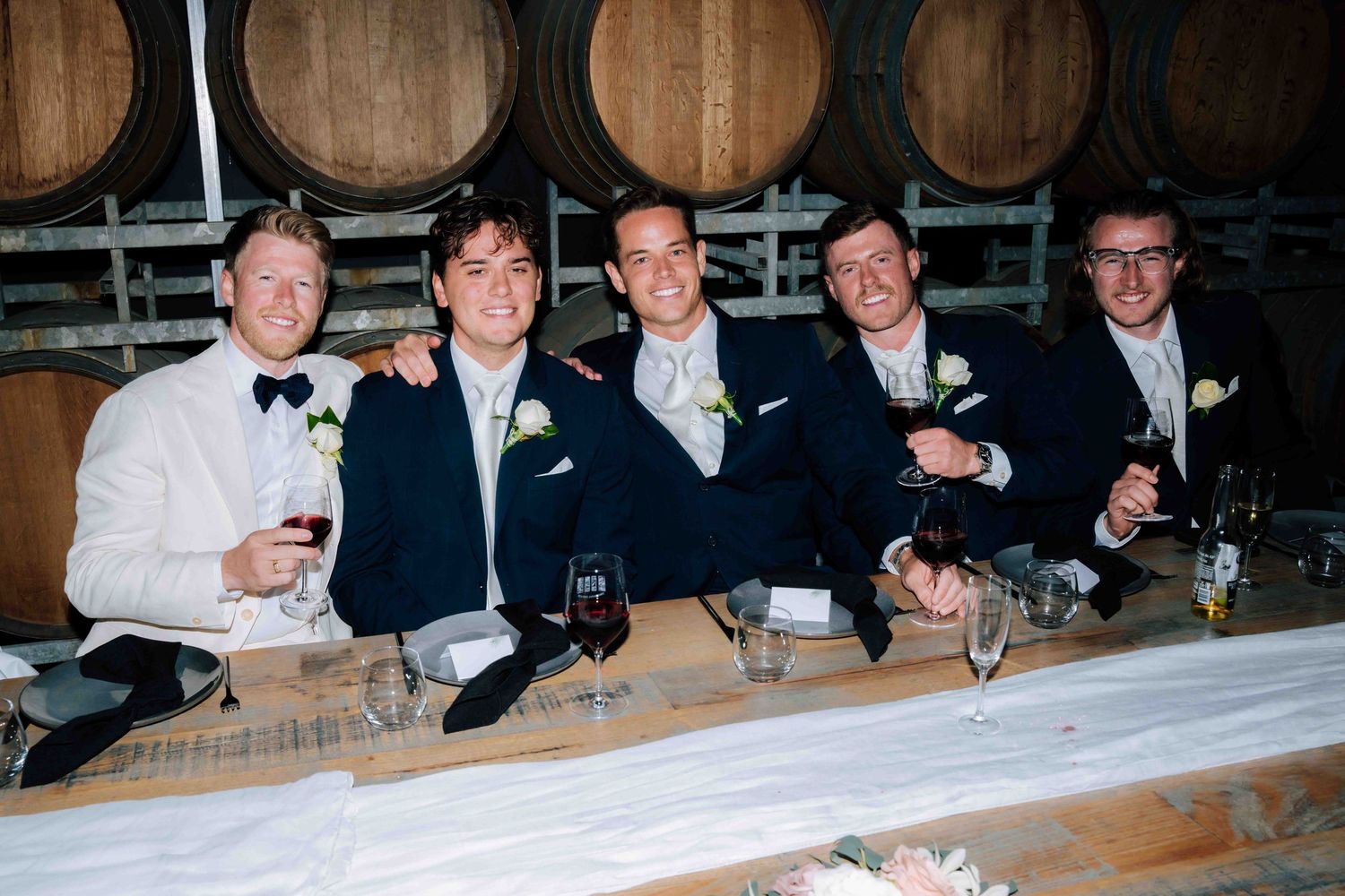 Five groomsmen smiling and toasting with wine glasses at a winery wedding reception.