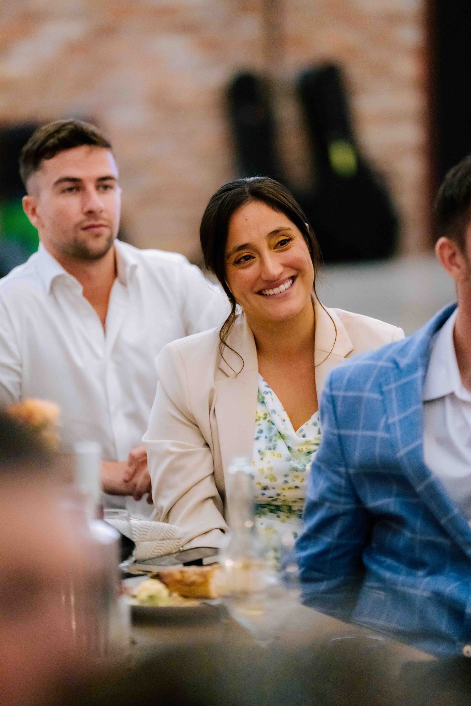 Smiling female wedding guest in cream blazer seated at reception dinner table.
