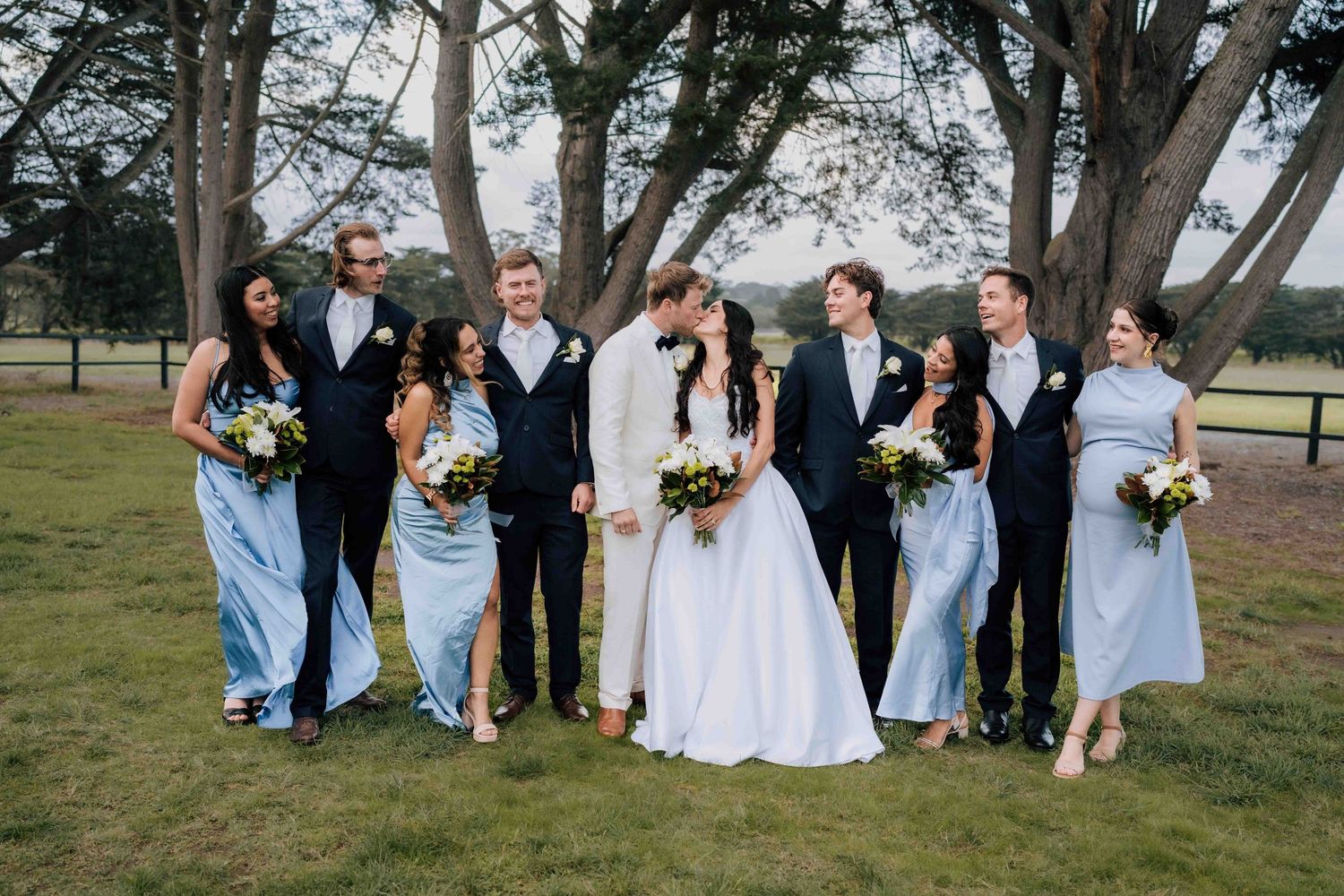 Wedding party poses outdoors with bride and groom kissing, bridesmaids in blue dresses and groomsmen in black suits.