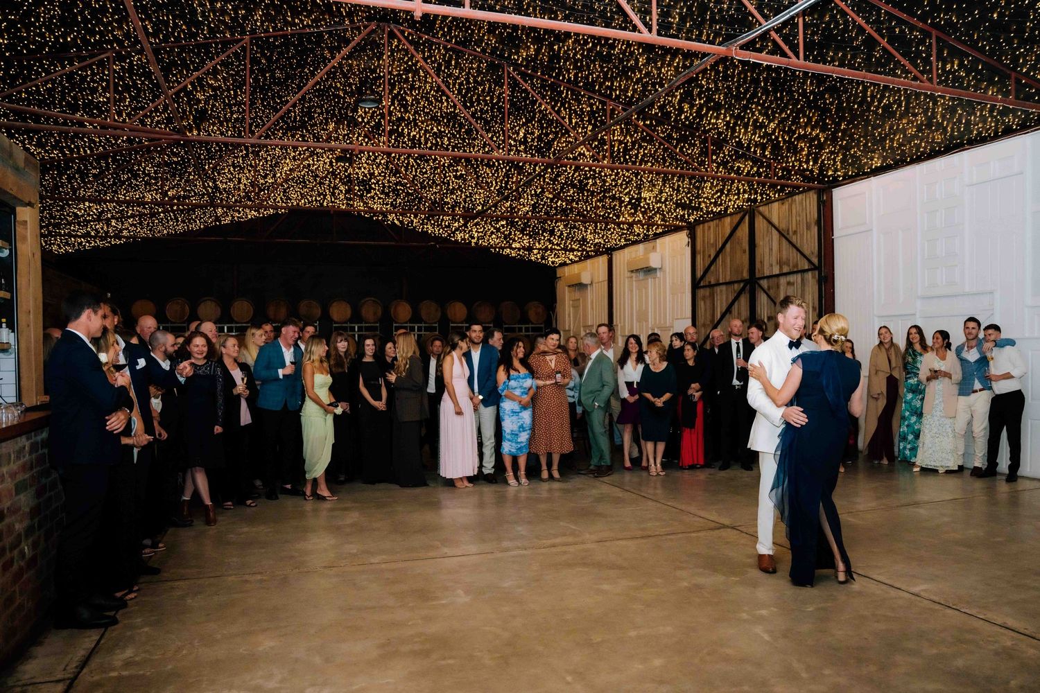 Couple sharing their first dance surrounded by wedding guests in a fairy light decorated barn.