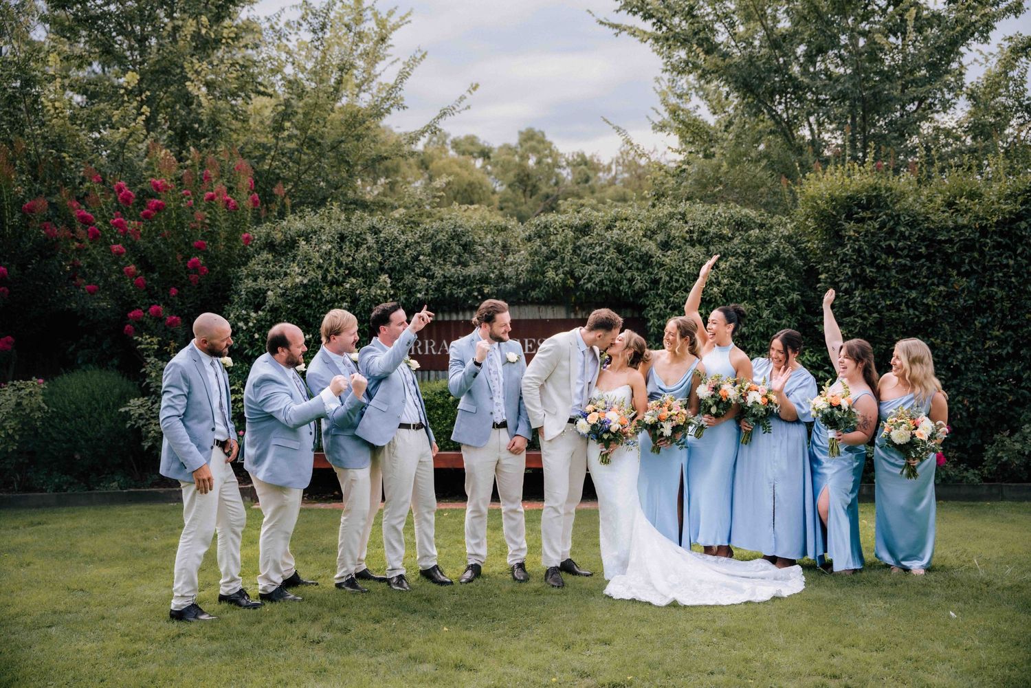 Joyful wedding party celebrates outdoors with bride and groom kissing surrounded by cheering guests.