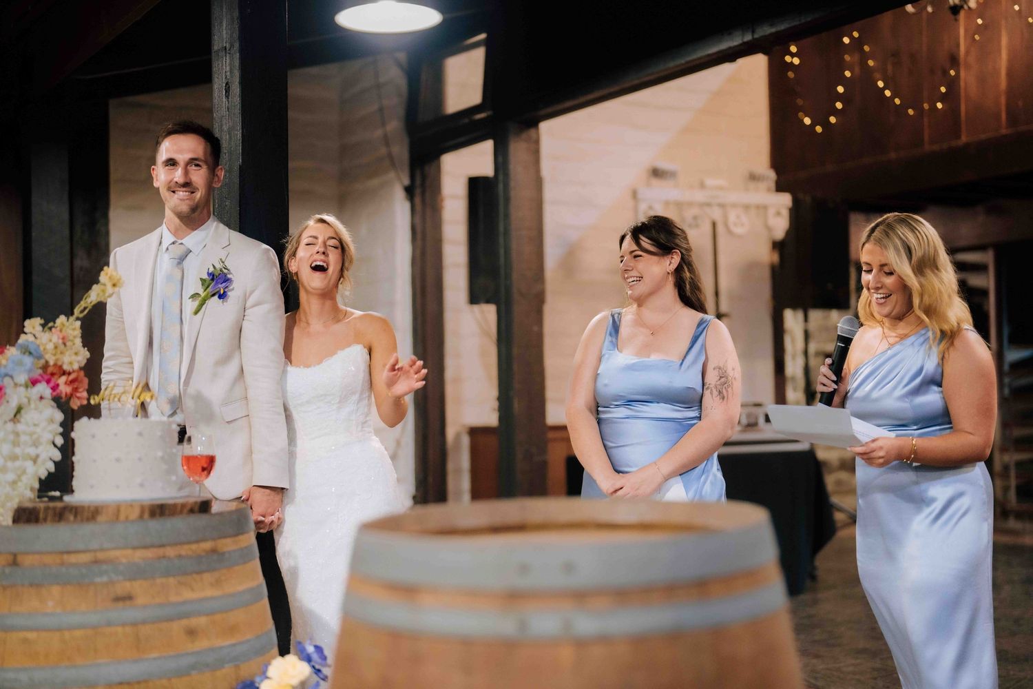 Bride and groom laugh with bridesmaids in blue dresses during wedding cake cutting ceremony.