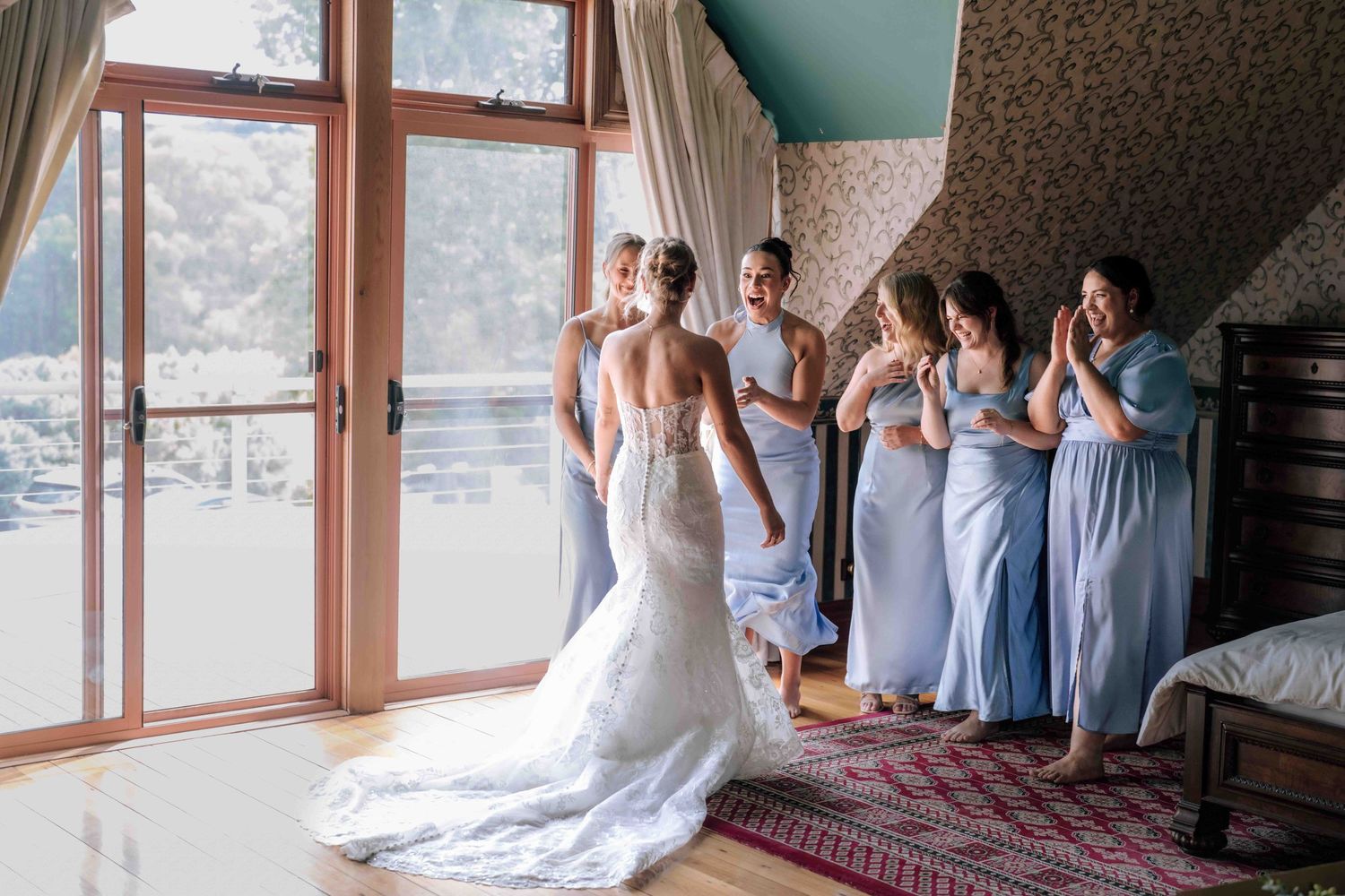 A bride in a lace wedding gown with a train stands before her bridesmaids in light blue dresses by large windows.
