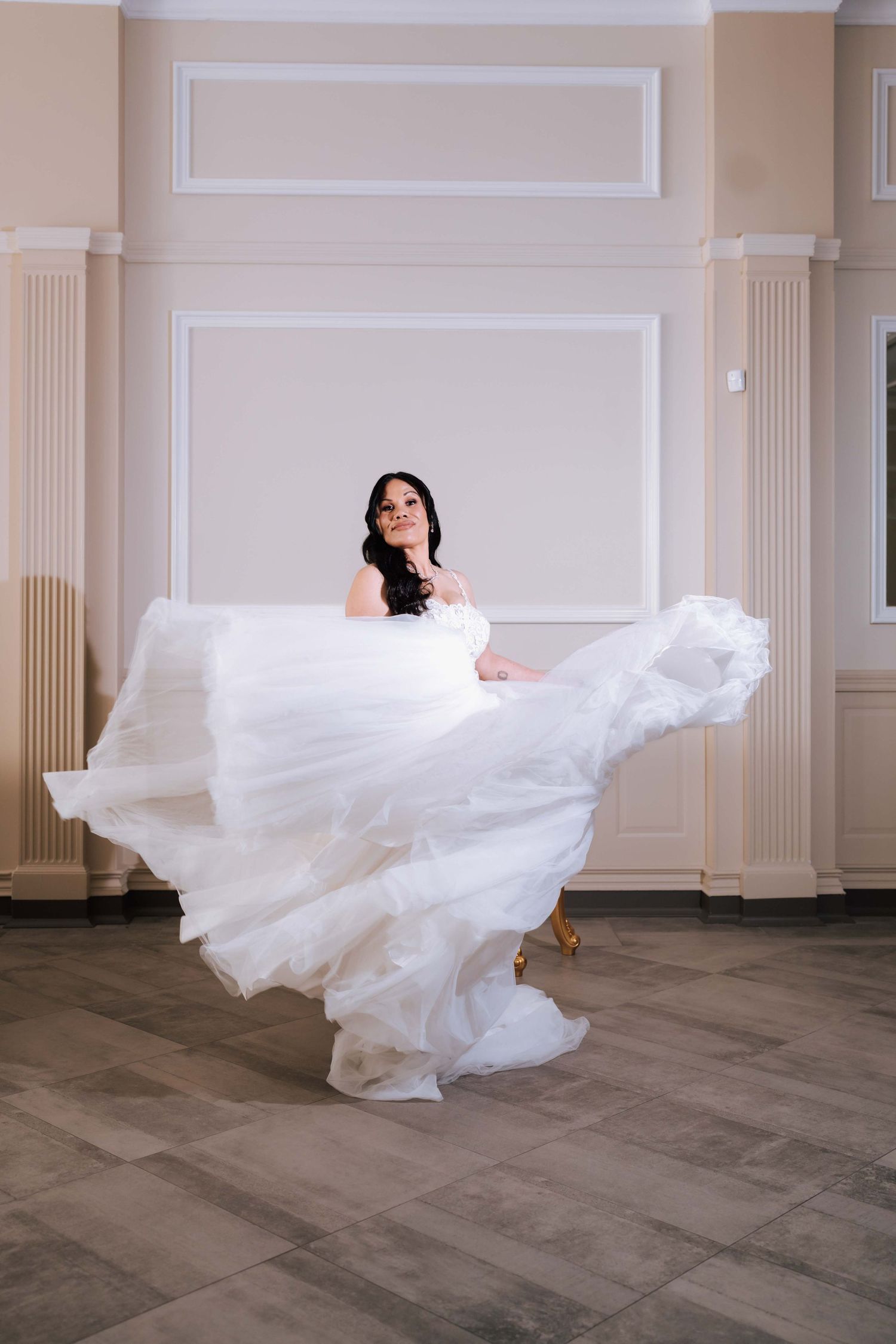 Bride in a flowing white ball gown twirling in an elegant room with classical white walls.