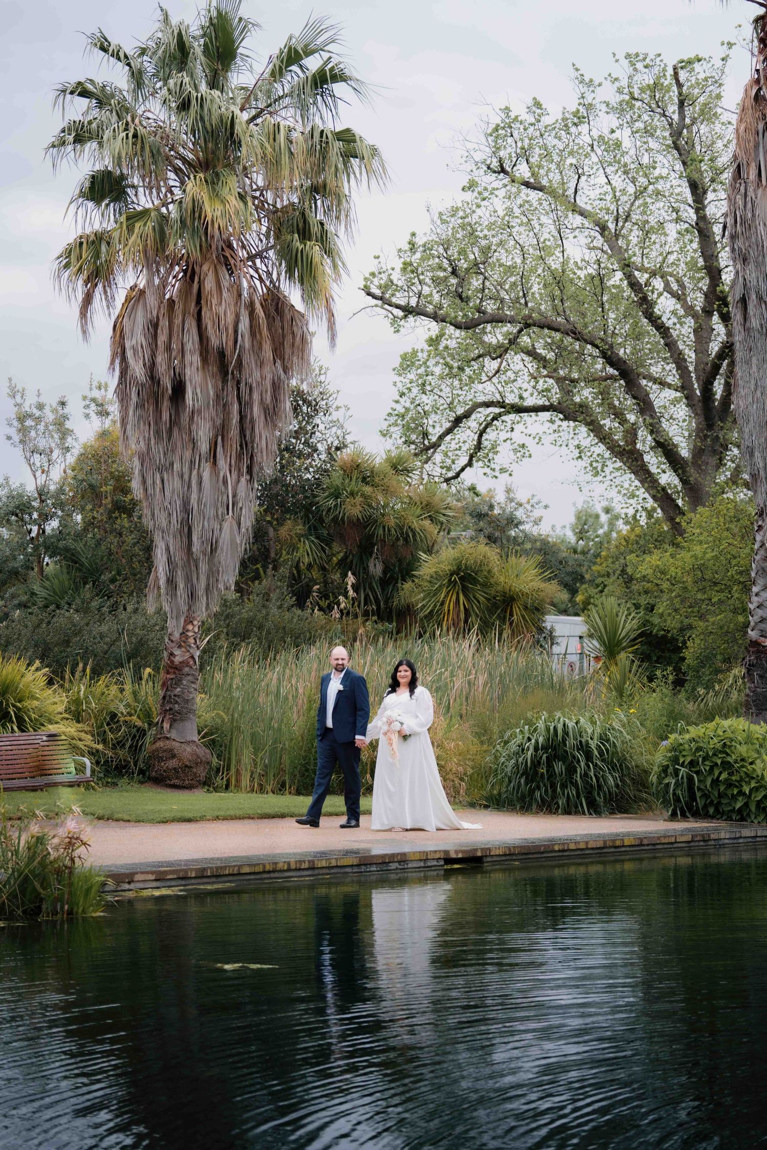 Bride and groom stand beside a reflective pond with palm trees and lush greenery on their wedding day.
