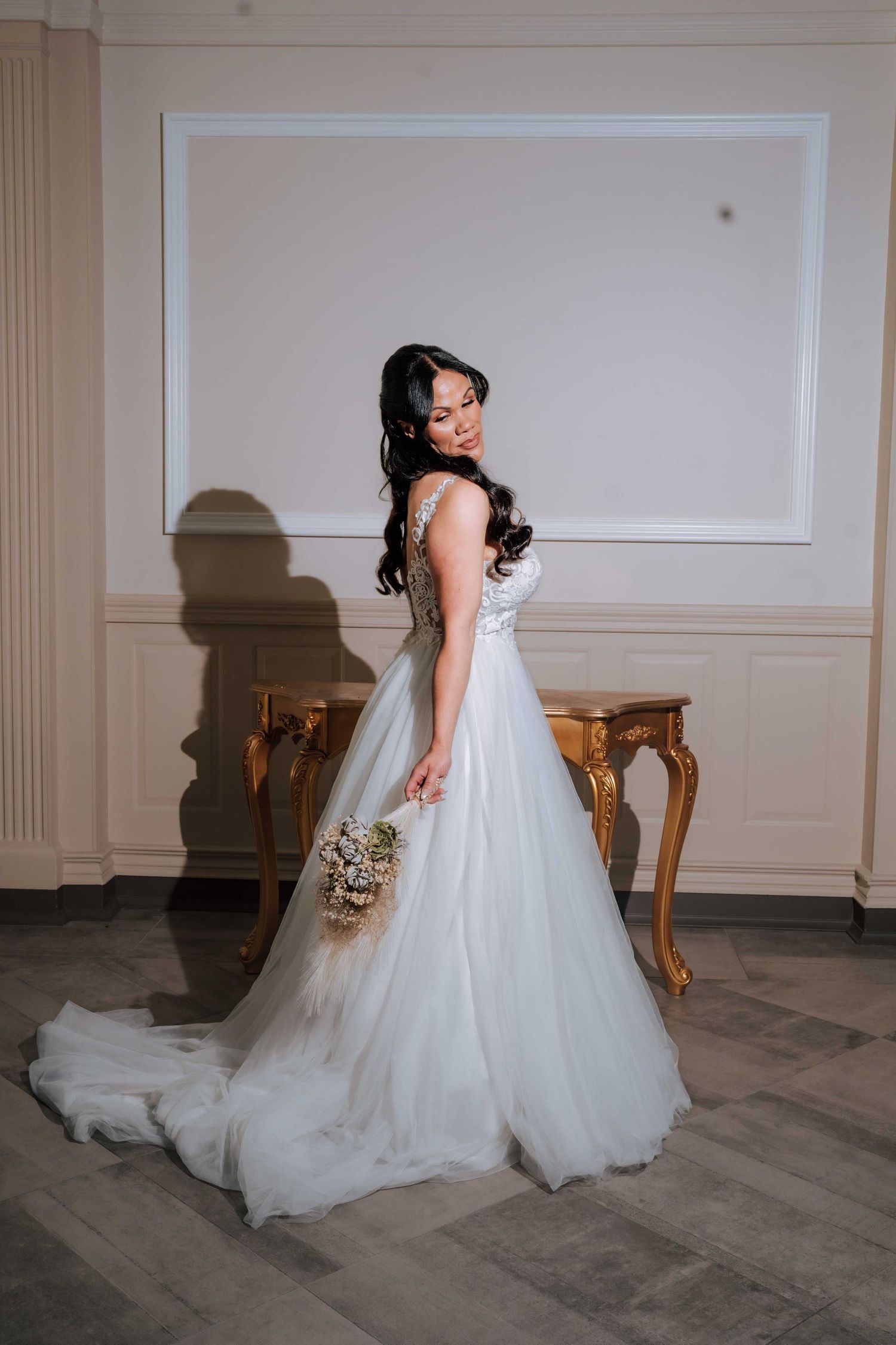 An elegant bride in a flowing white tulle wedding gown holds a bouquet in a classic interior room.