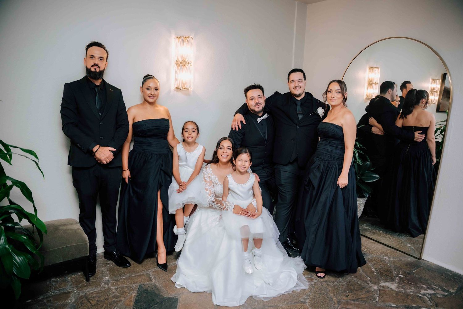 Wedding party in elegant black attire poses with bride and flower girls in white dresses indoors.