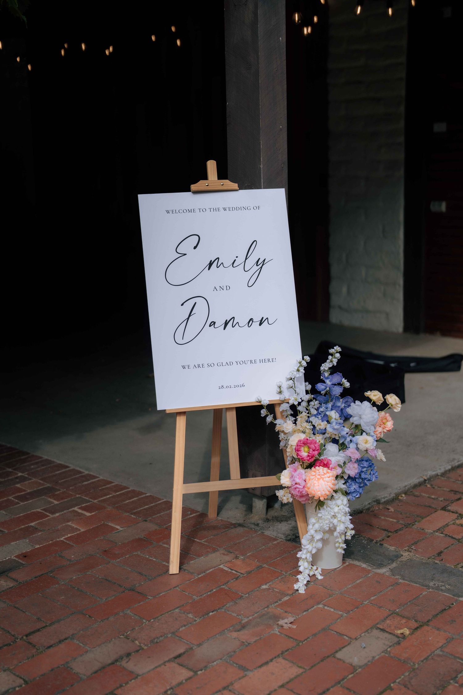 Elegant wedding welcome sign for Emily and Damon on a wooden easel with colorful floral arrangement on brick pathway.