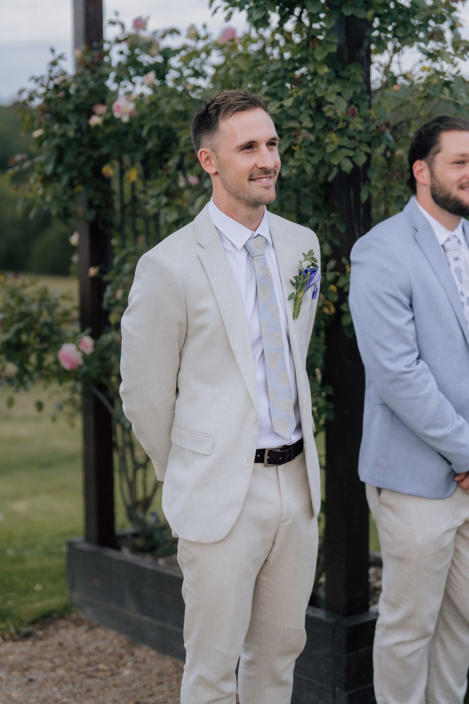 Groom in beige suit with floral boutonniere smiles during outdoor wedding ceremony beside groomsman.