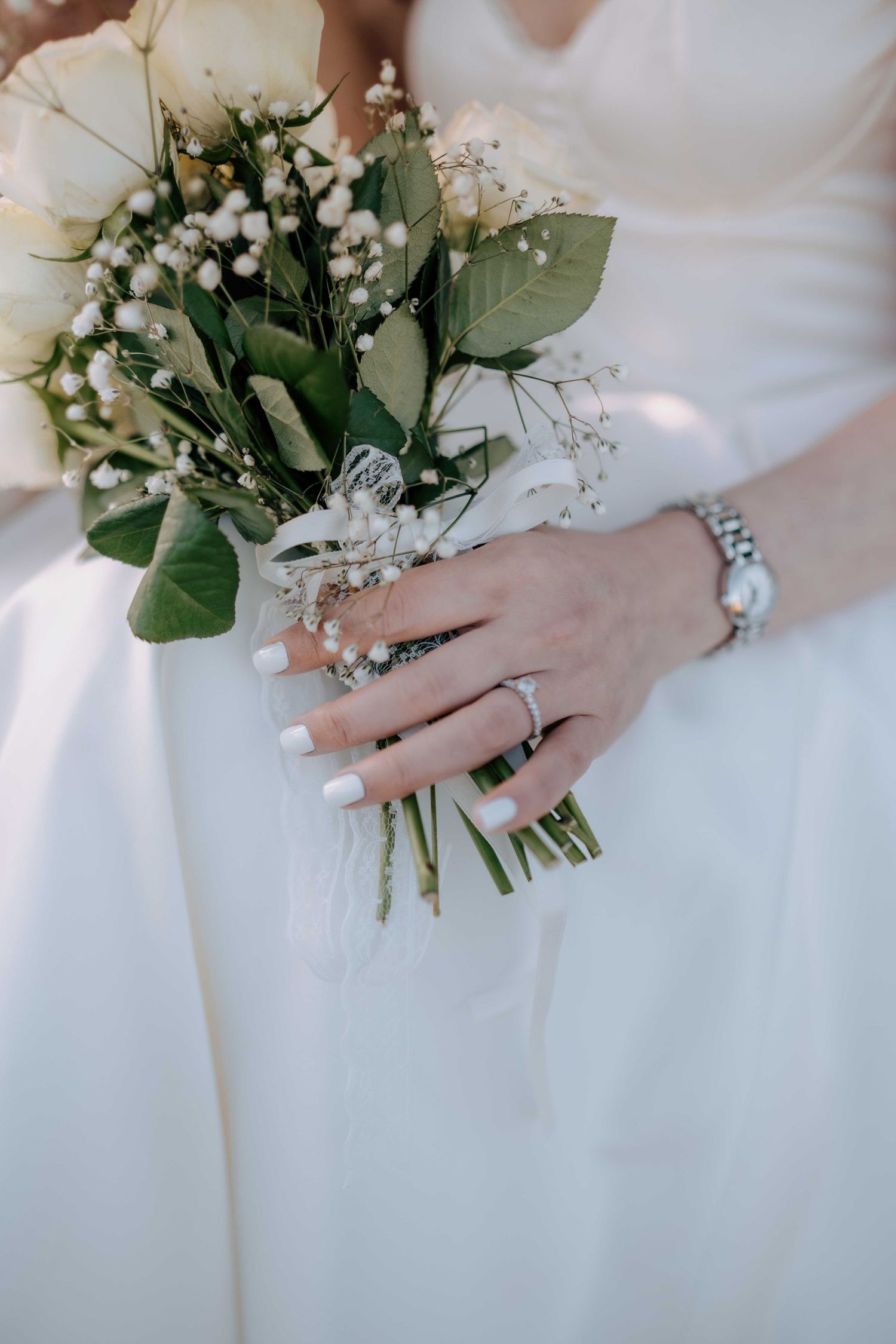 Bride holds white rose bouquet with baby's breath, showing diamond ring and silver bracelet on wedding day.