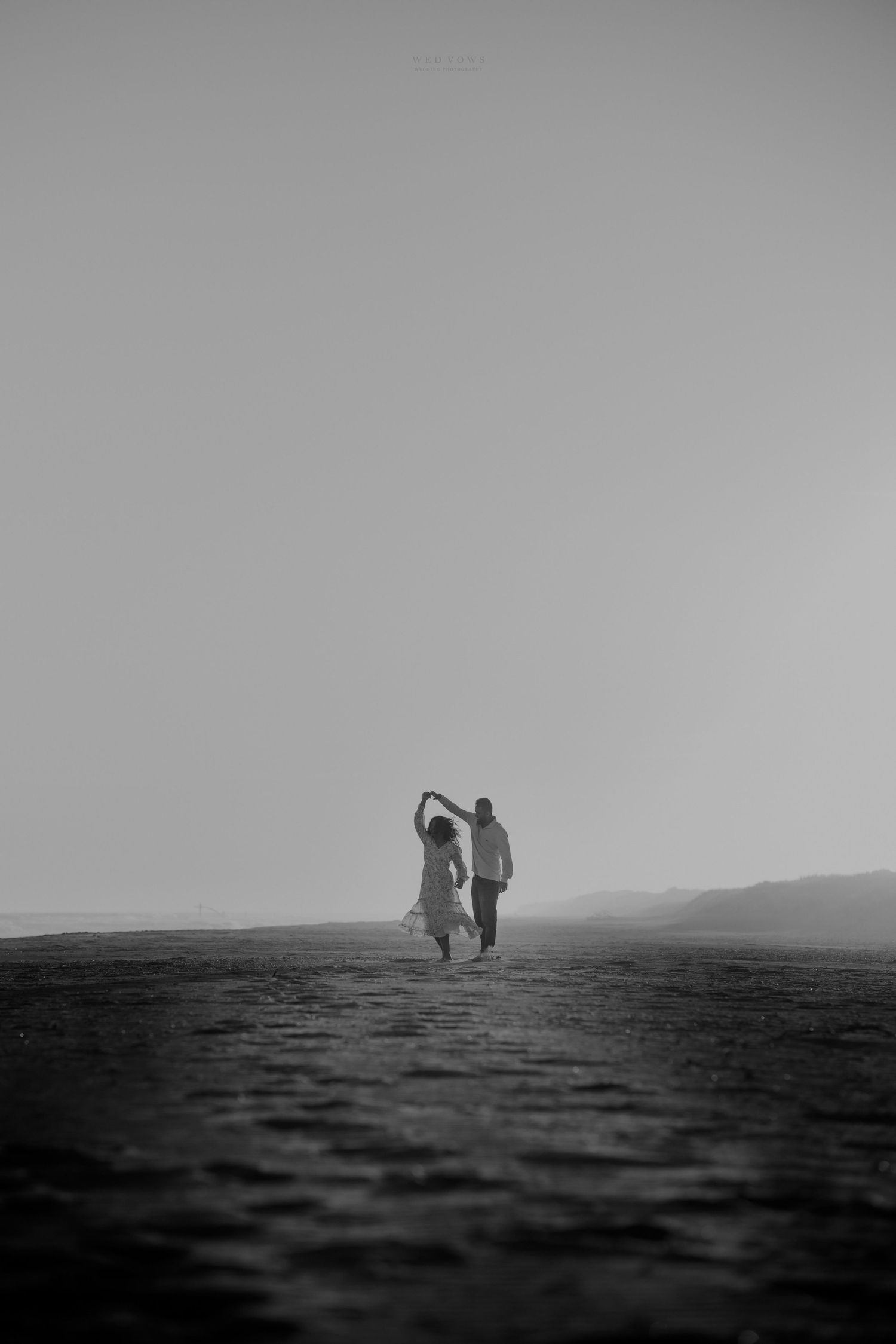 Black and white photo of a couple holding hands and dancing on a misty beach at sunset.