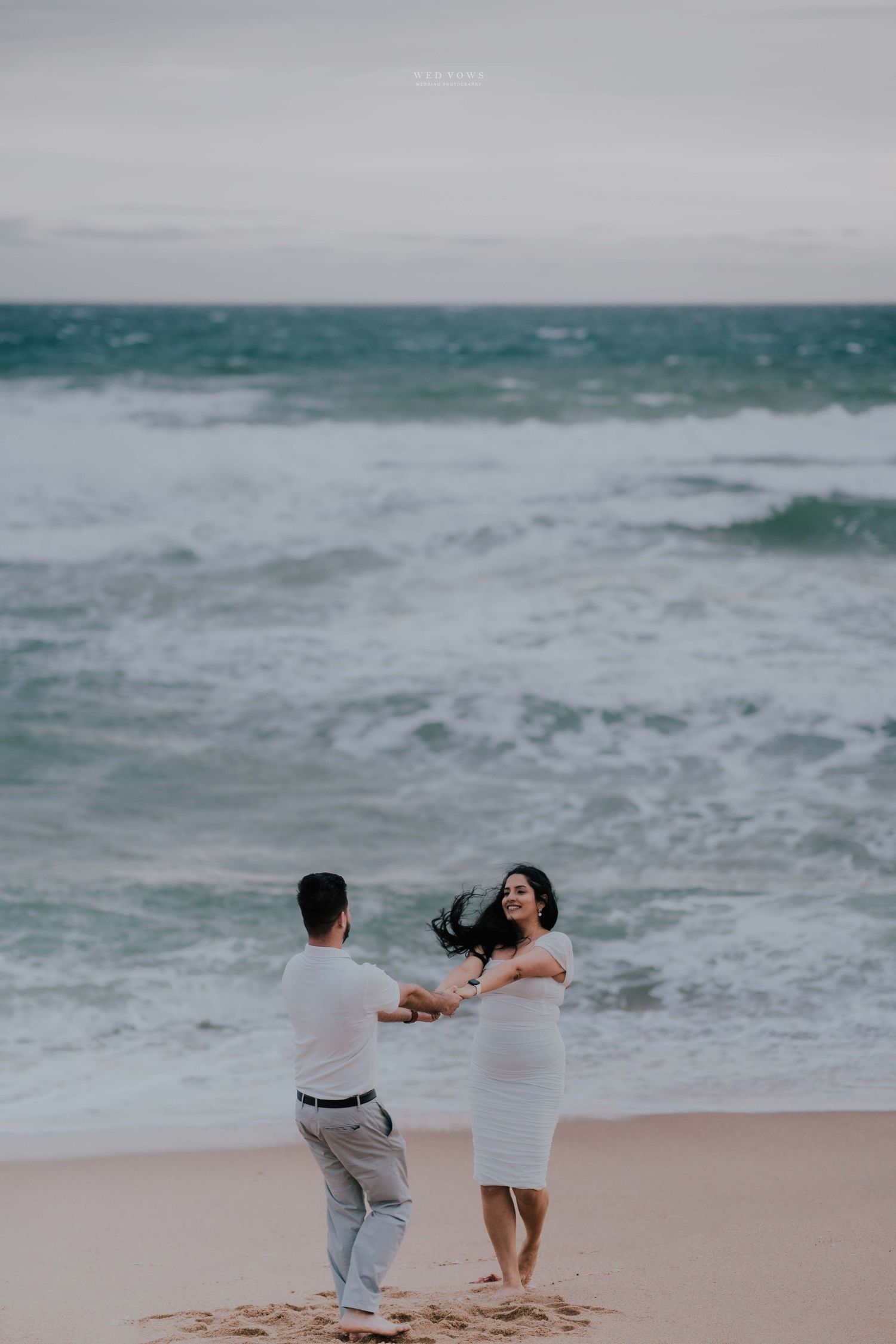 Couple dancing joyfully on a windy beach with dramatic ocean waves in the background.